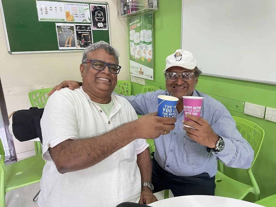 Two smiling men clink paper cups in a bright green room with chairs and posters on the wall.