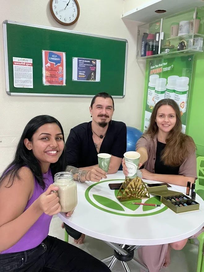 Three people sitting at a round table in a room, smiling and holding paper cups, with a jar of smoothie or milkshake in front of the woman on the left, and a box of nail polish or essential oils on the table.