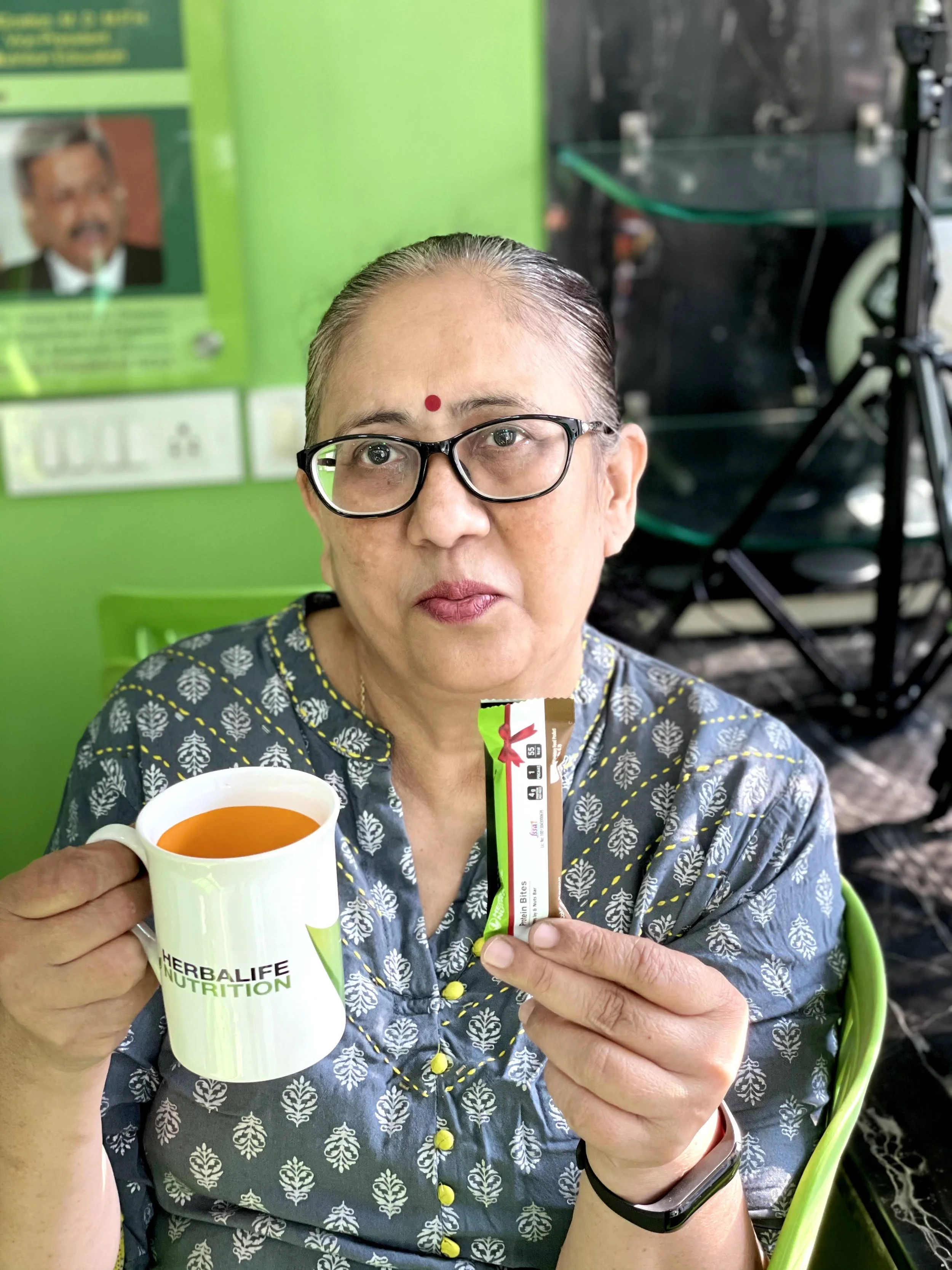 An older woman with glasses, wearing a patterned shirt, holding a white mug with Indian tea and a herbal supplement packet. She has a red bindi on her forehead and is sitting in a setting with a green wall in the background.