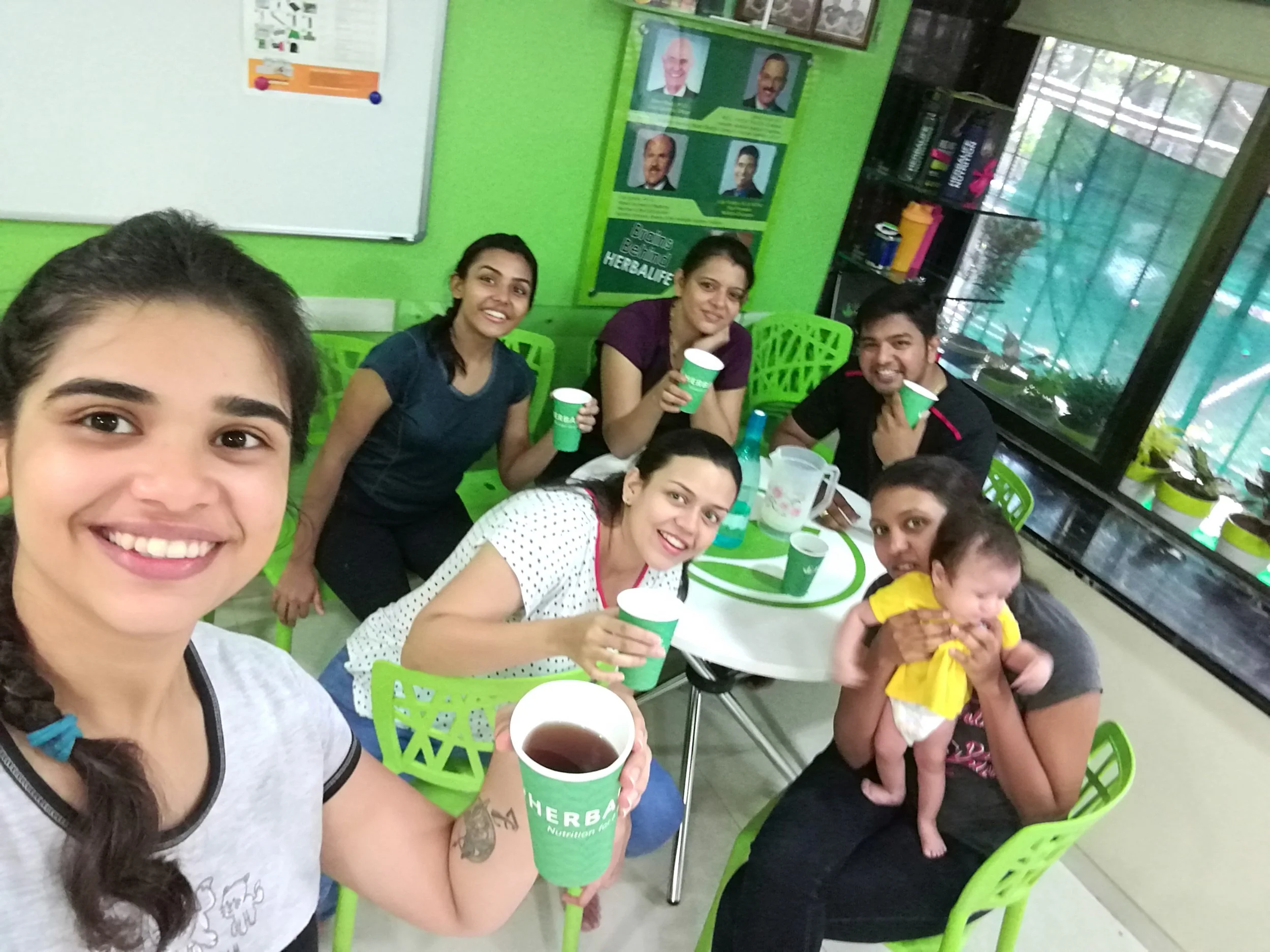 Group of six people, five women and one man, sitting around a green and white table with cups, smiling and taking a selfie. One woman is holding a cup of tea or coffee, and another woman is holding a baby; all appear to be enjoying a casual gathering