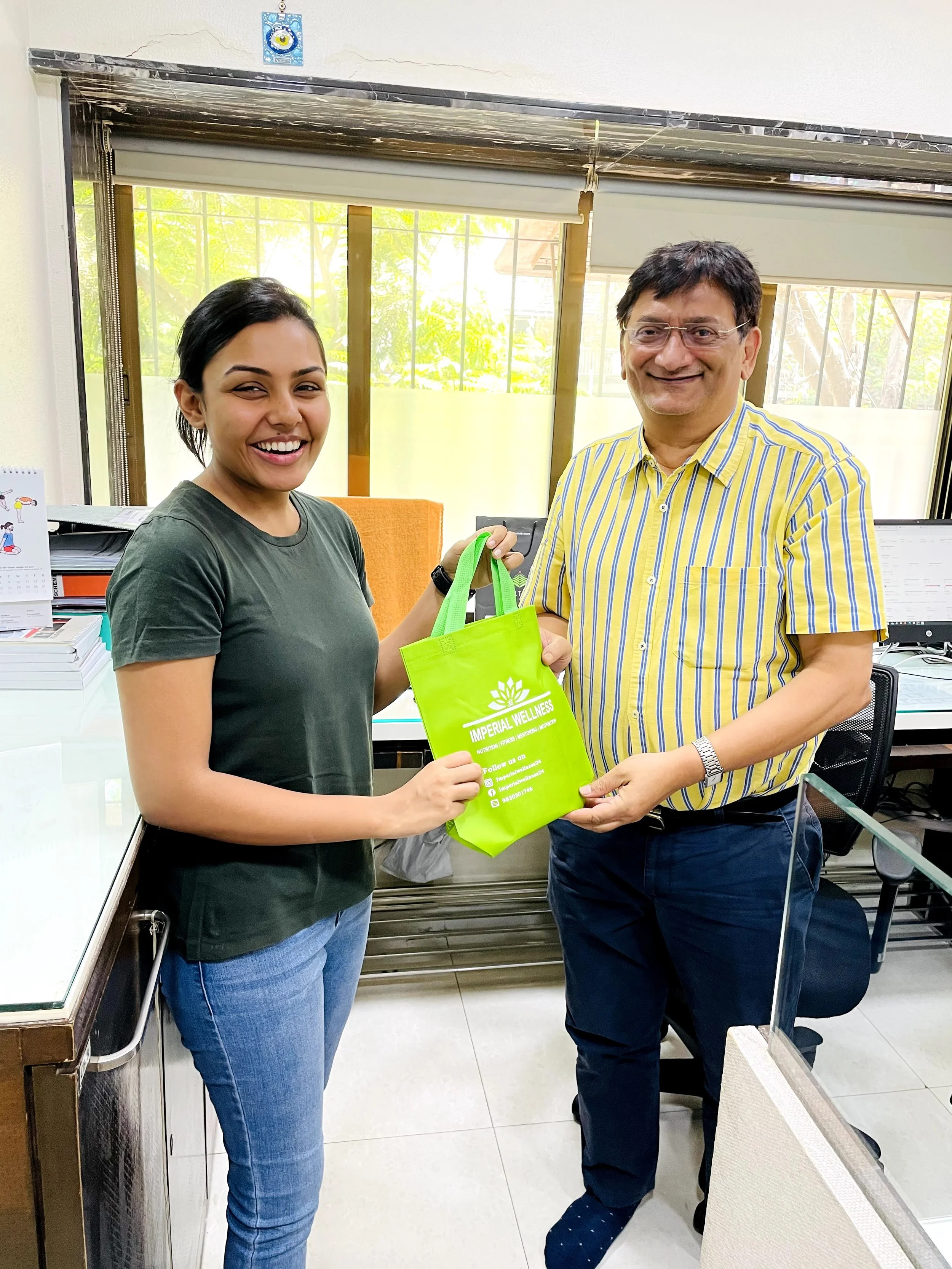 Woman and man exchanging a green tote bag with 'Imperial Wellness' printed on it in an office setting.