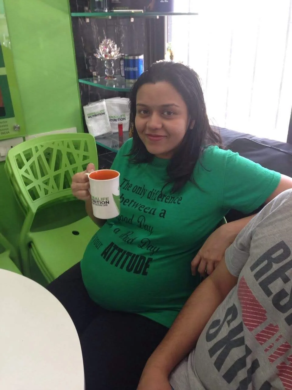 A woman with dark hair, wearing a green T-shirt with black text, is sitting at a table, holding a white mug with a red interior and the Herbalife Nutrition logo. She is smiling and sitting next to a person wearing a gray T-shirt. The background inclu
