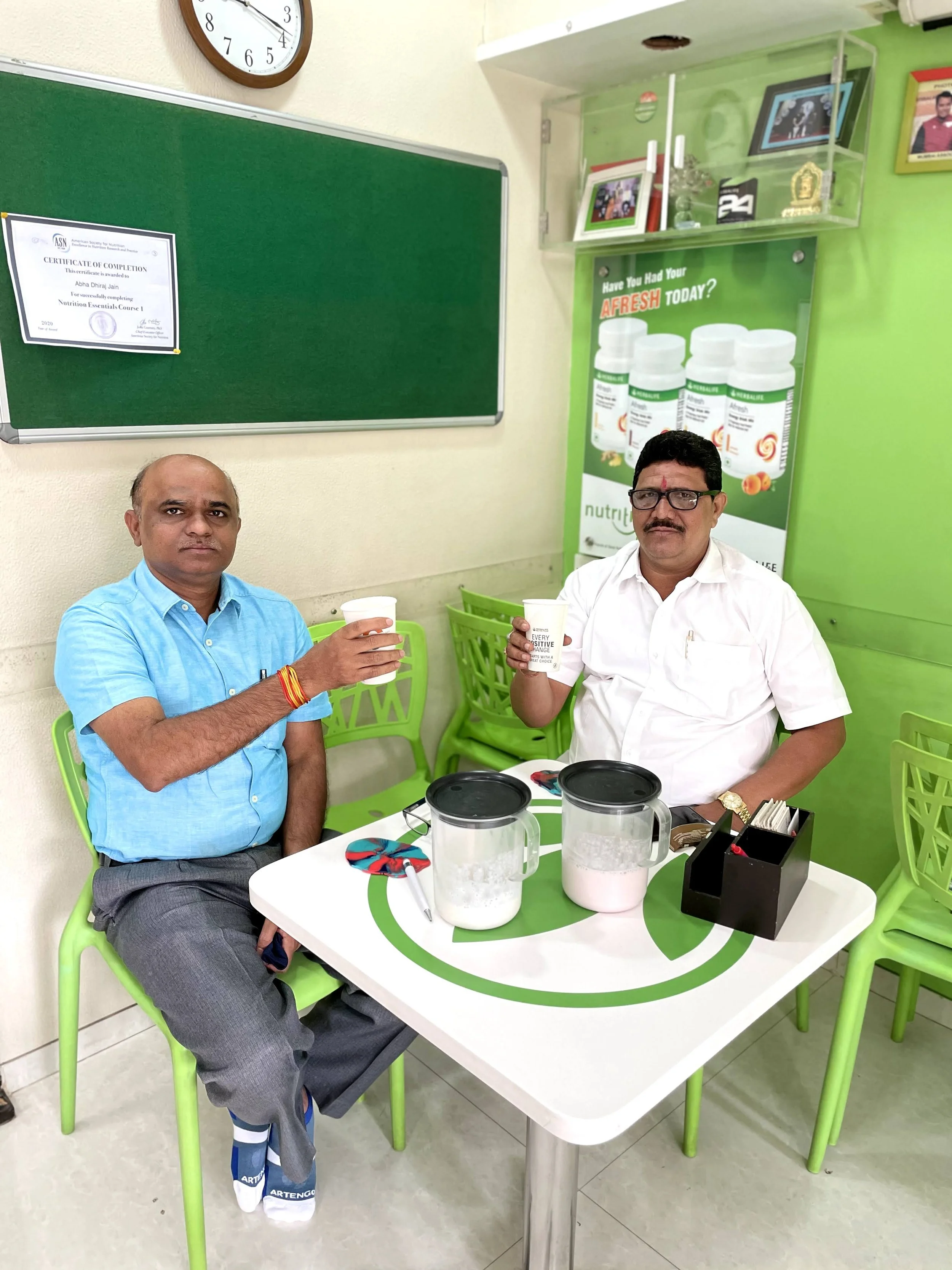 Two men sitting at a white table, holding white cups, in a green-themed room with a blackboard and a poster advertising herbal supplements. The man on the left is dressed in a blue shirt and gray trousers, while the man on the right is wearing a whit