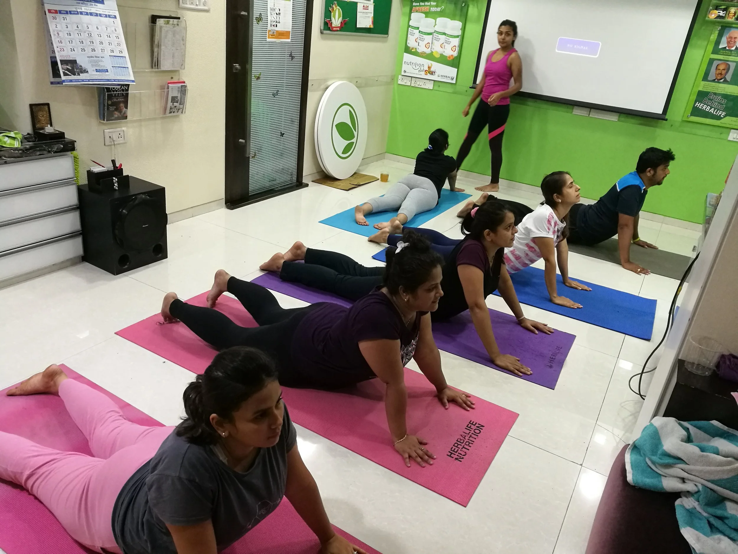 A group of women participating in a yoga class in a room with green walls, on colorful mats, guided by an instructor standing in front of a whiteboard.