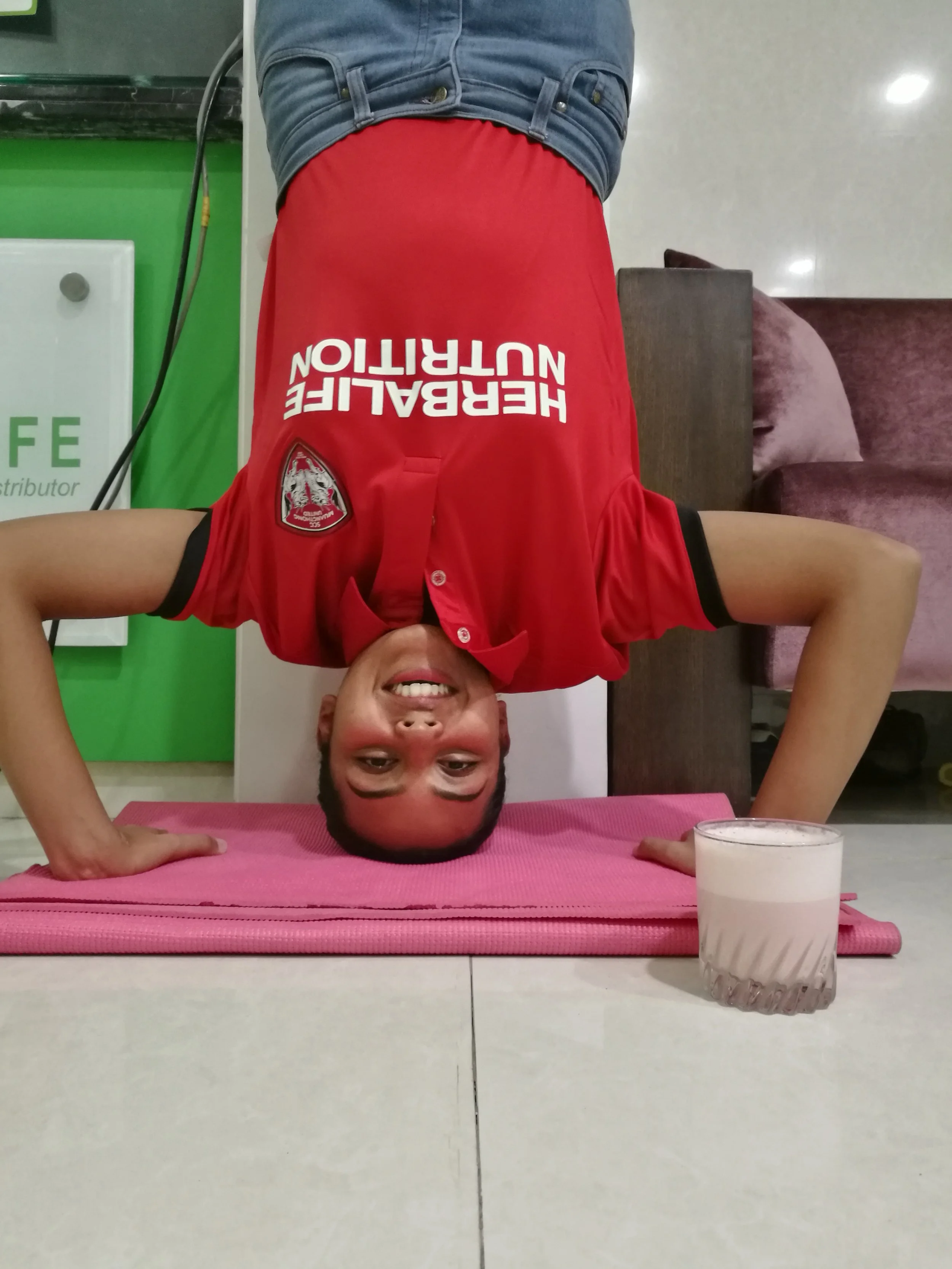 A person doing a headstand on a pink yoga mat, smiling, with a glass of milk nearby. The person is wearing a red shirt with white text and blue jeans, indoors with a green wall and brown/gray furniture in the background.