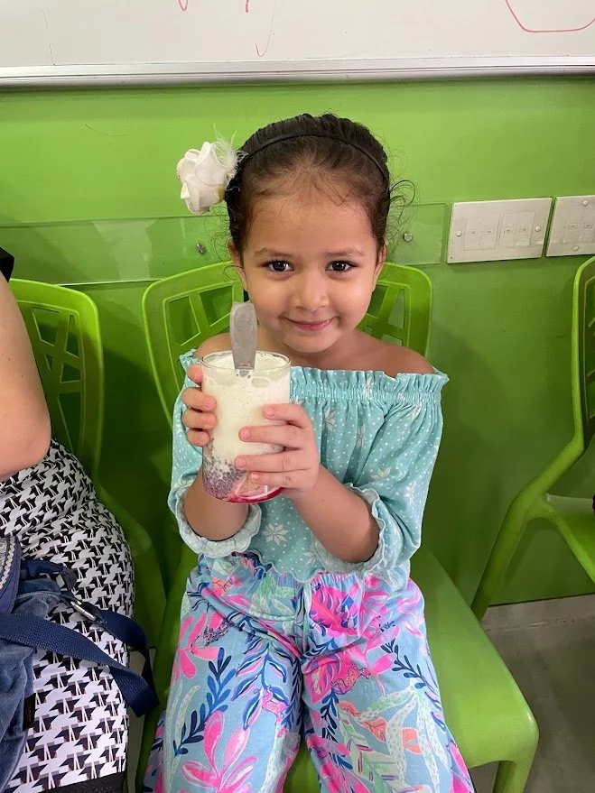 A young girl with a white hair bow, wearing a light blue off-the-shoulder top and colorful pants, sitting on a green chair, holding a glass of what appears to be a pink and white layered beverage with a spoon inside, in a room with green walls and el