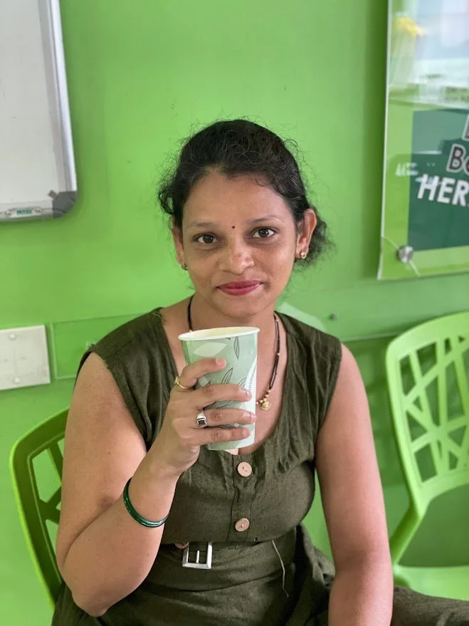A woman sitting on a green chair holding a paper cup, with a bright green wall and a partially visible sign behind her.