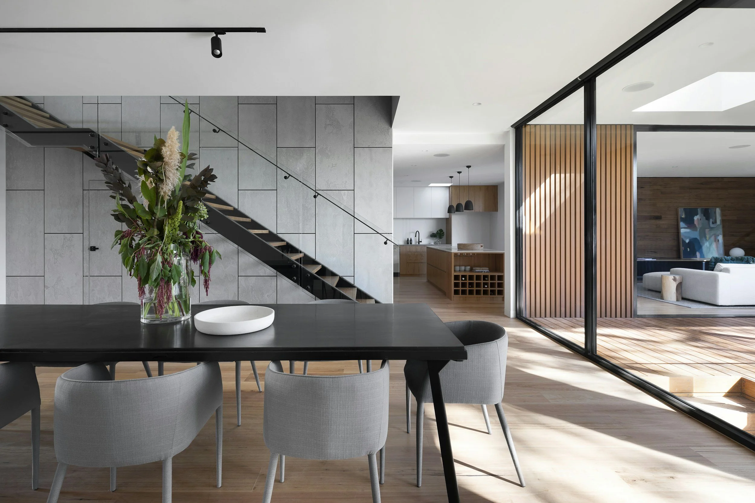 Modern dining area with black table and gray chairs, glass vase with flowers, staircase with metal railing, open kitchen with island, pendant lights, and sliding glass doors leading to living room.