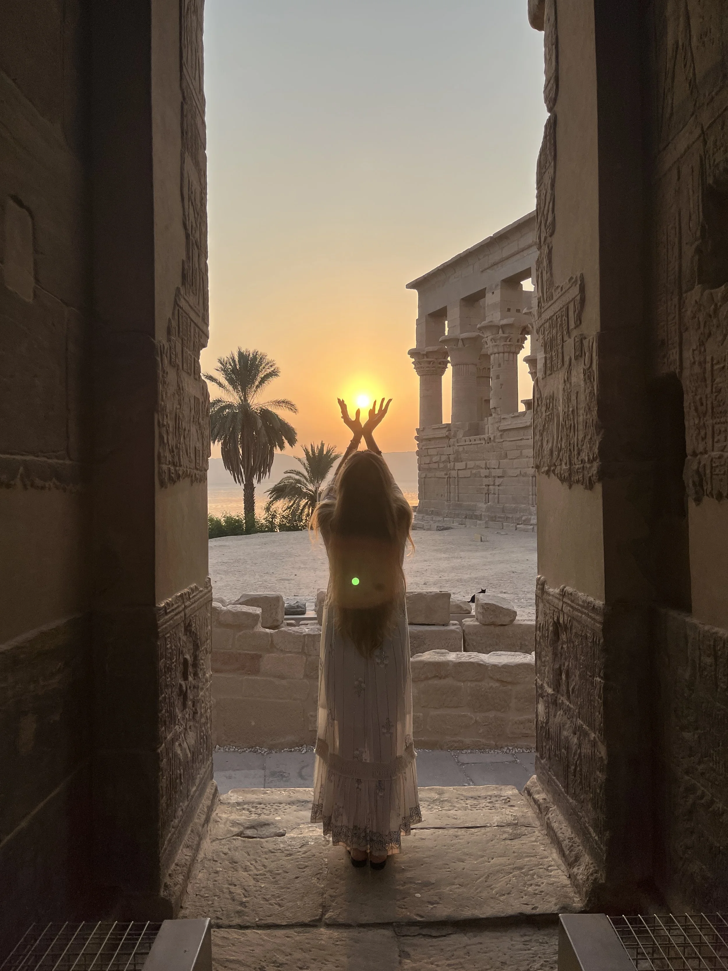 Sabina standing in a temple doorway at sunset with hands raised toward the sun at Philae Temple in Egypt during a sacred pilgrimage