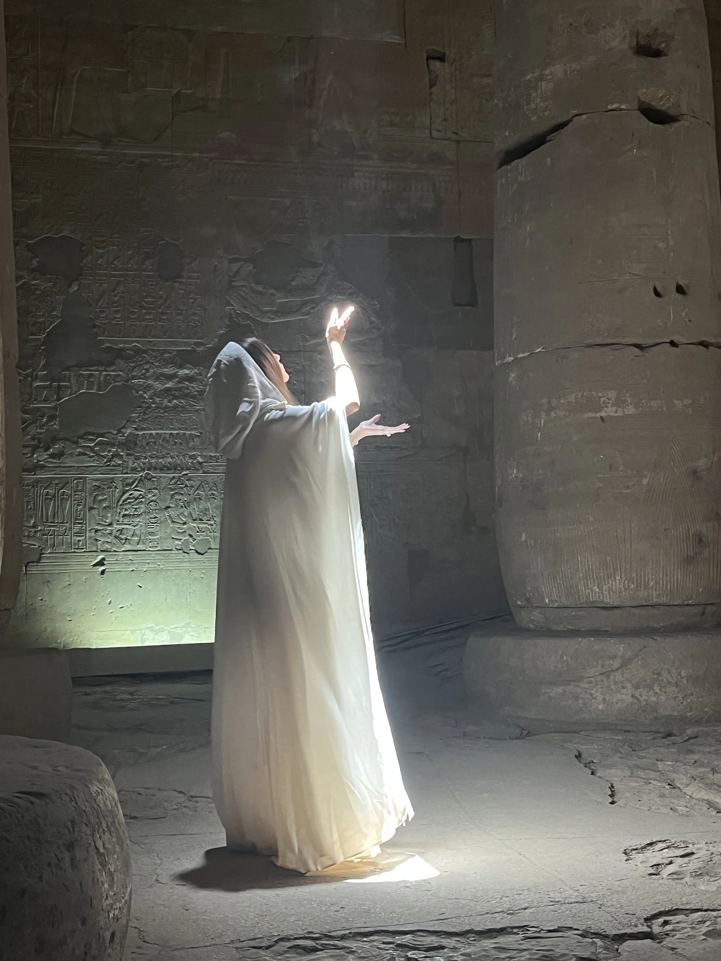 Sabina in white hooded dress with hand raised and glowing light inside an ancient Egyptian temple during a sacred ceremony
