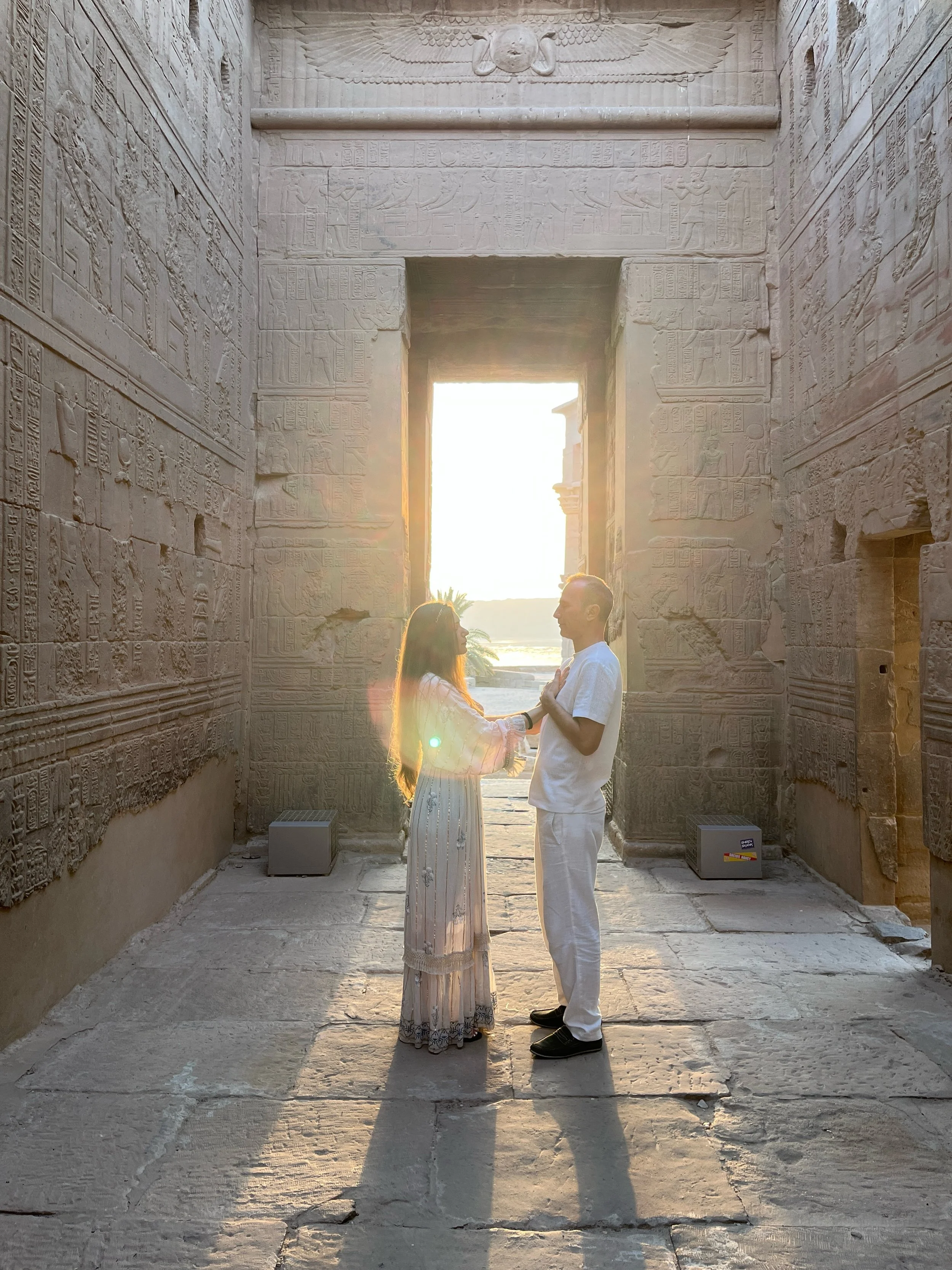 Sabina and Leo in prayer hands facing each other inside an ancient Egyptian temple corridor with sunlight behind them