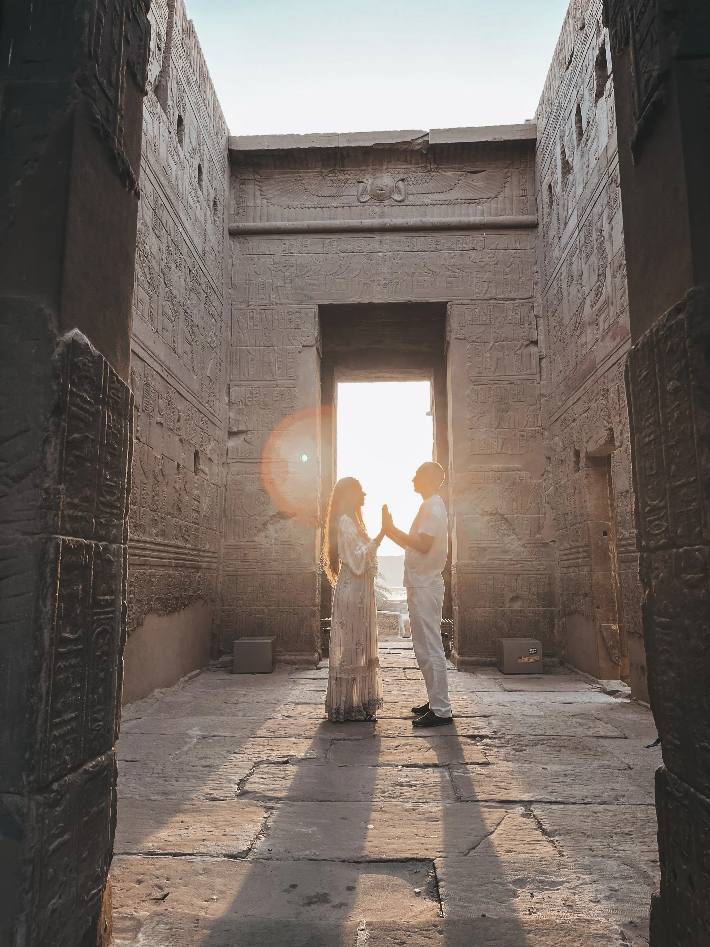 Sabina and Leo facing each other in prayer hands inside an ancient Egyptian temple corridor with golden sunlight streaming through the doorway during sacred ceremony