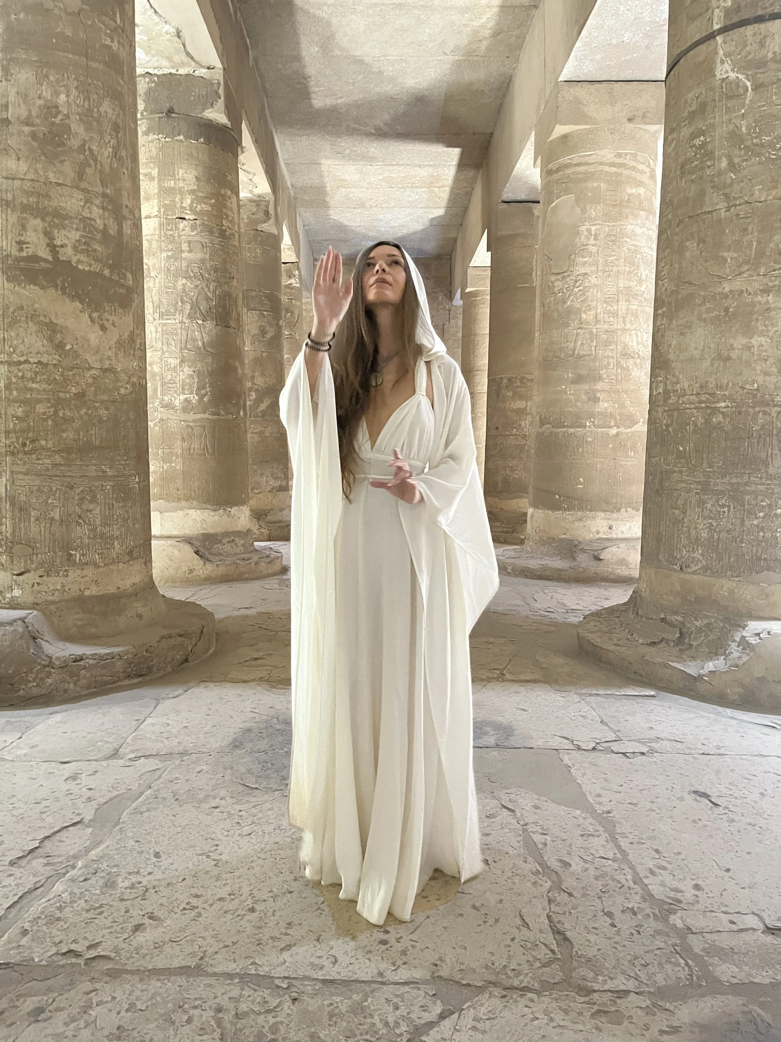 Sabina in white dress with one hand raised in sacred gesture and light codes channeling inside an ancient Egyptian temple with carved columns and hieroglyphs