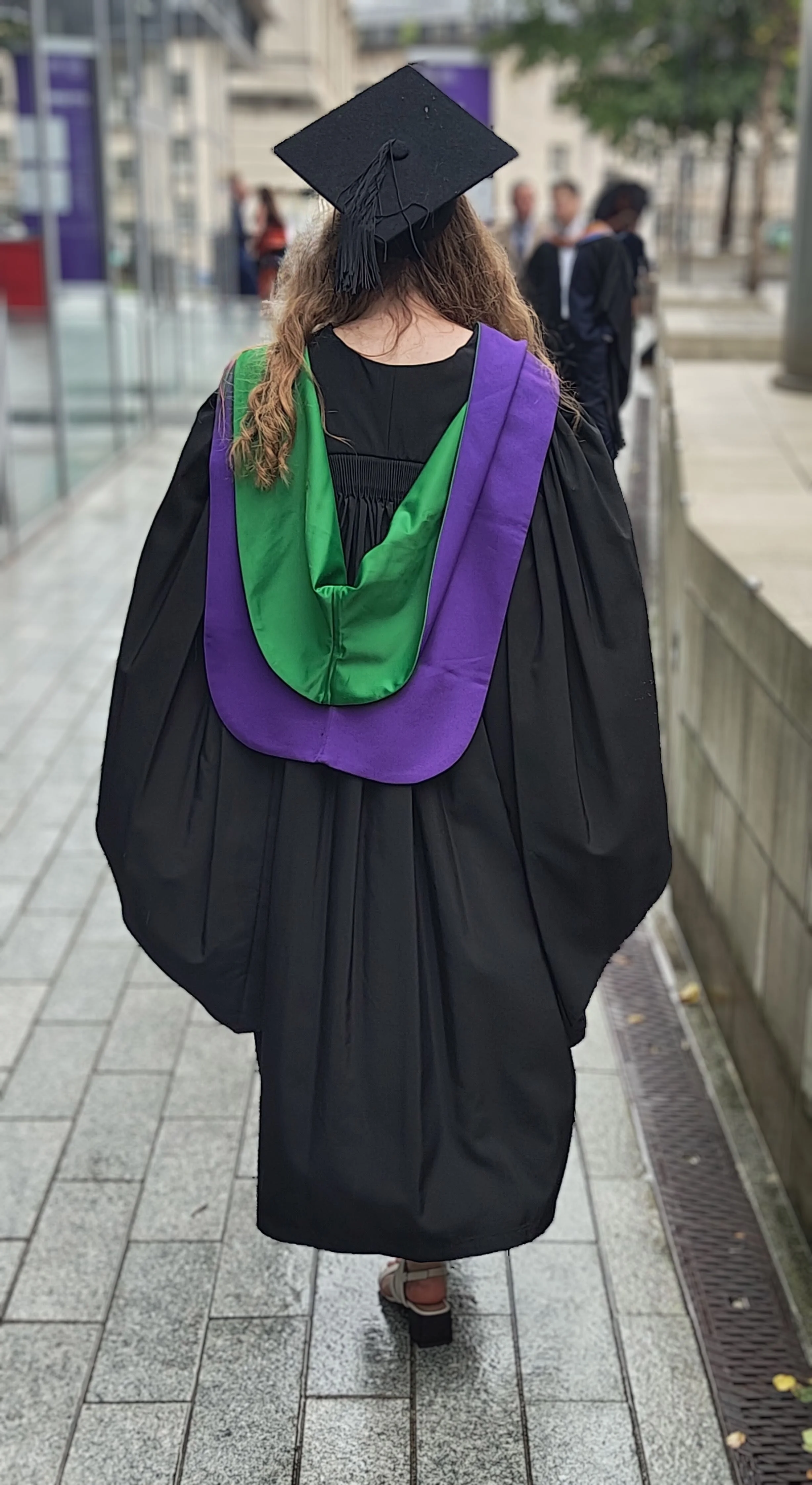 Woman wearing a black graduation gown and cap, walking on a city sidewalk during daytime.