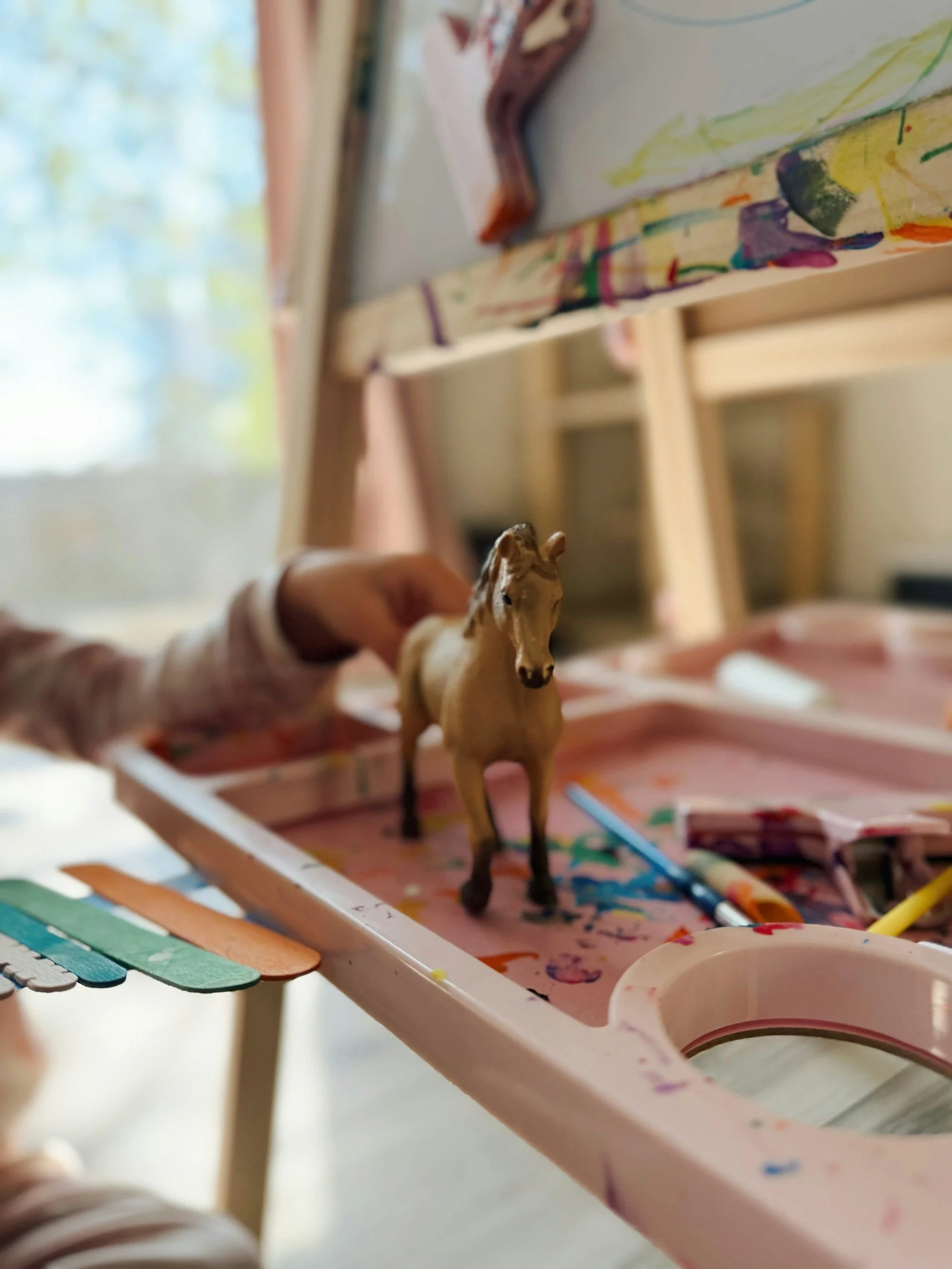 A small toy horse placed on a pink paint-splattered palette with paintbrushes and colorful sticks nearby, with a blurred background of a window and blinds.