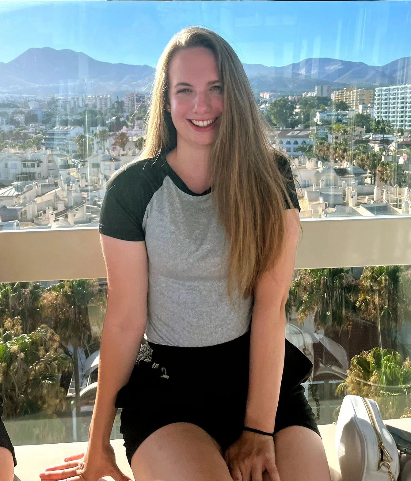 A woman with long blonde hair smiling, sitting on a ledge with a cityscape and mountains in the background.