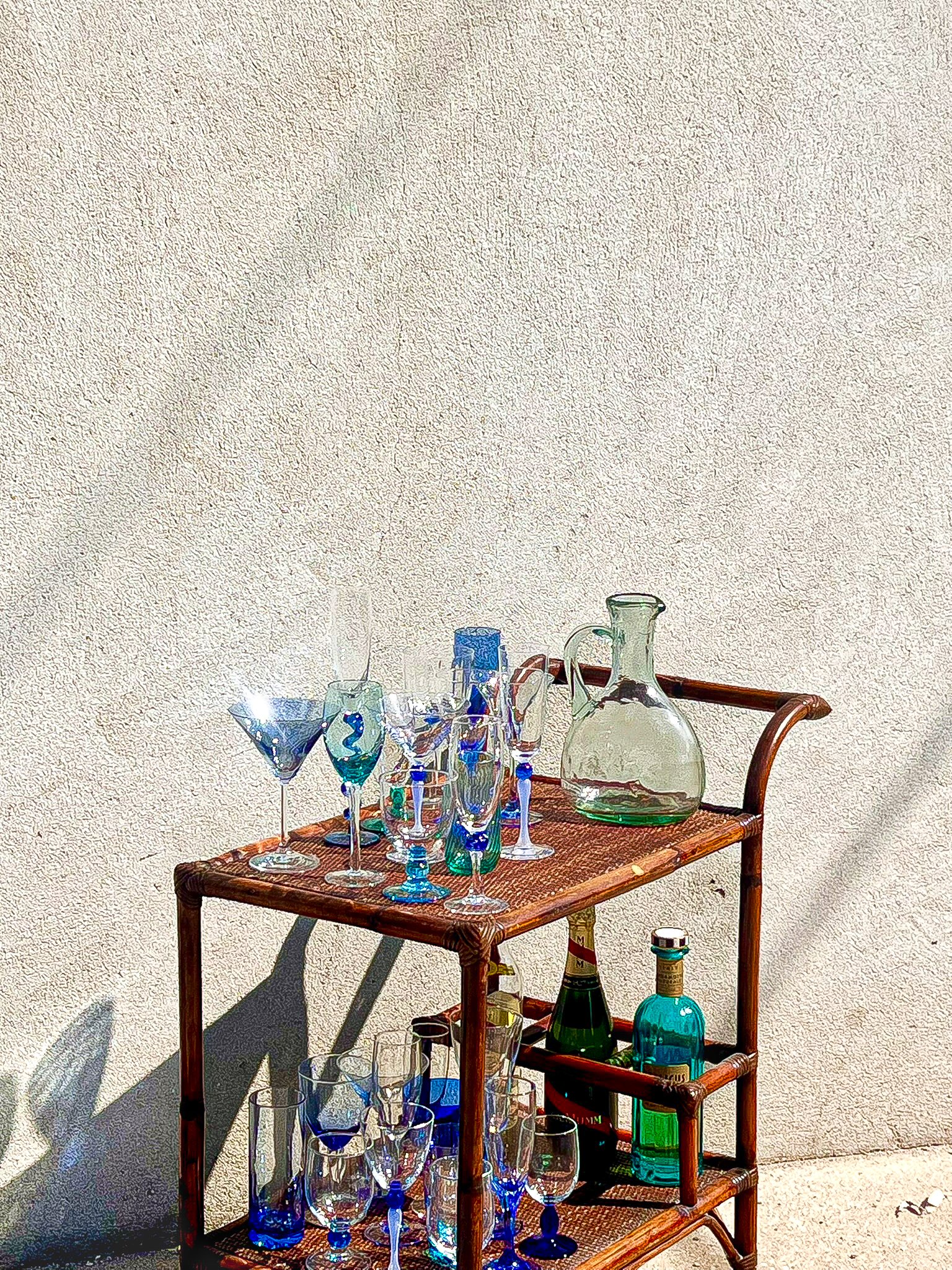Location de mobilier pour évènement. Dessert chariot vintage en osier avec des verres colorés et des bouteilles. Verres à pieds bleus et verts pour table colorée. location de verres colorés pour évènement, mariage et anniversaire à Paris et Ile de Fr