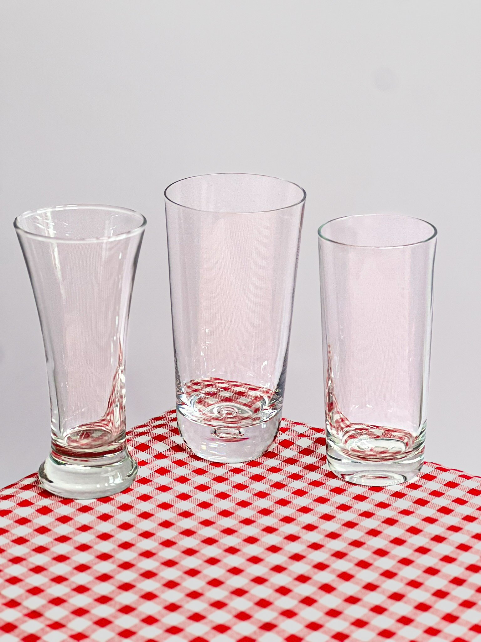 Verres à cocktails dépareillés chinés de formes différentes sur une table à motif vichy rouge et blanc. Verres dépareillés à louer pour évènements à Paris. Location verres pour mariage et anniversaire