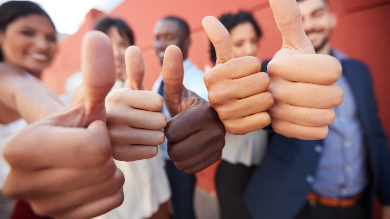 Eine Gruppe von Menschen zeigt Daumen hoch vor einer roten Wand, wobei die Kamera den Fokus auf die Daumen legt.