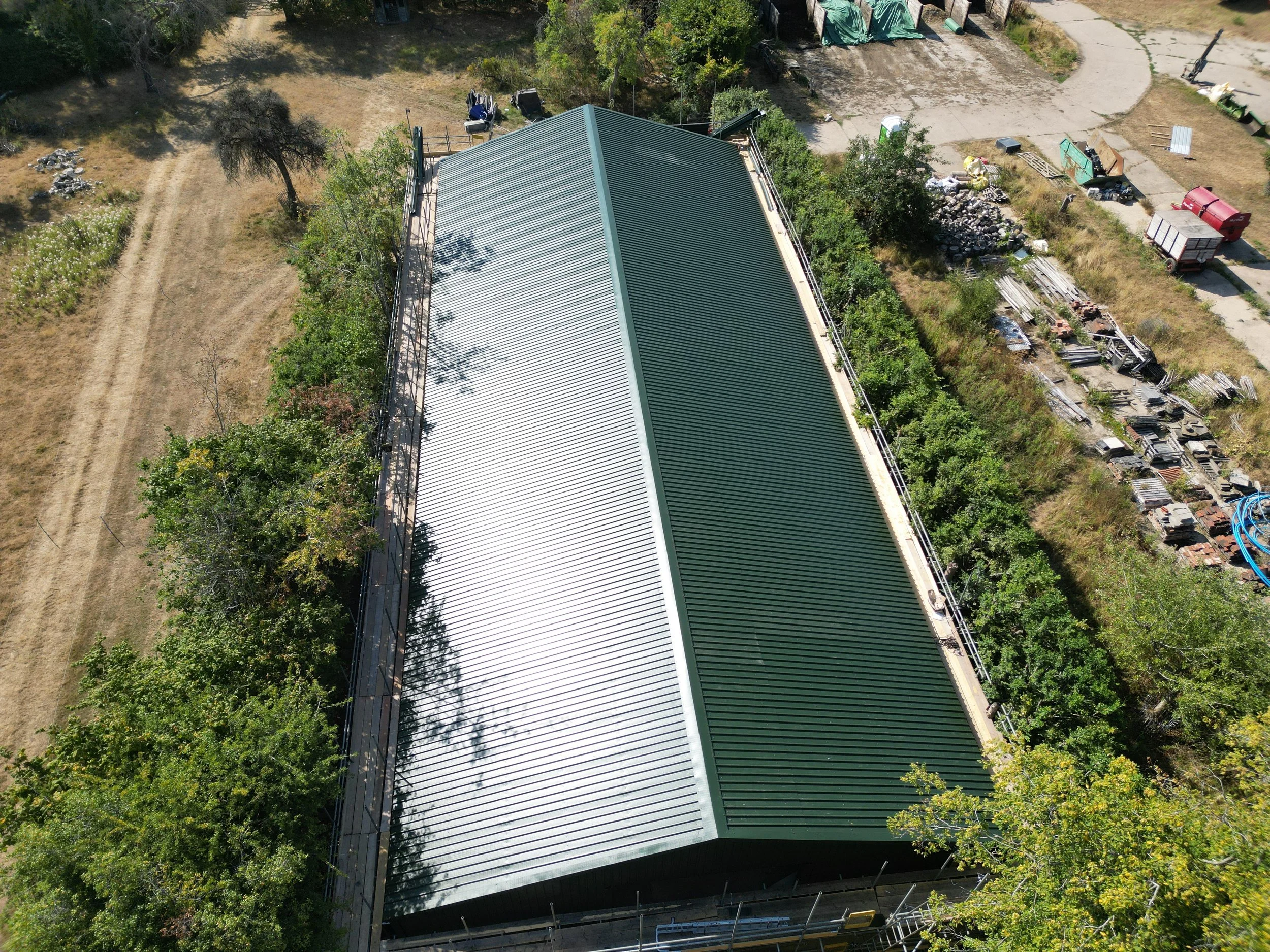 An aerial view of a large, rectangular building with a metal roof, surrounded by trees and a yard. There are construction materials, equipment, and vehicles nearby, as well as a dirt path and a paved area with various objects and debris.
