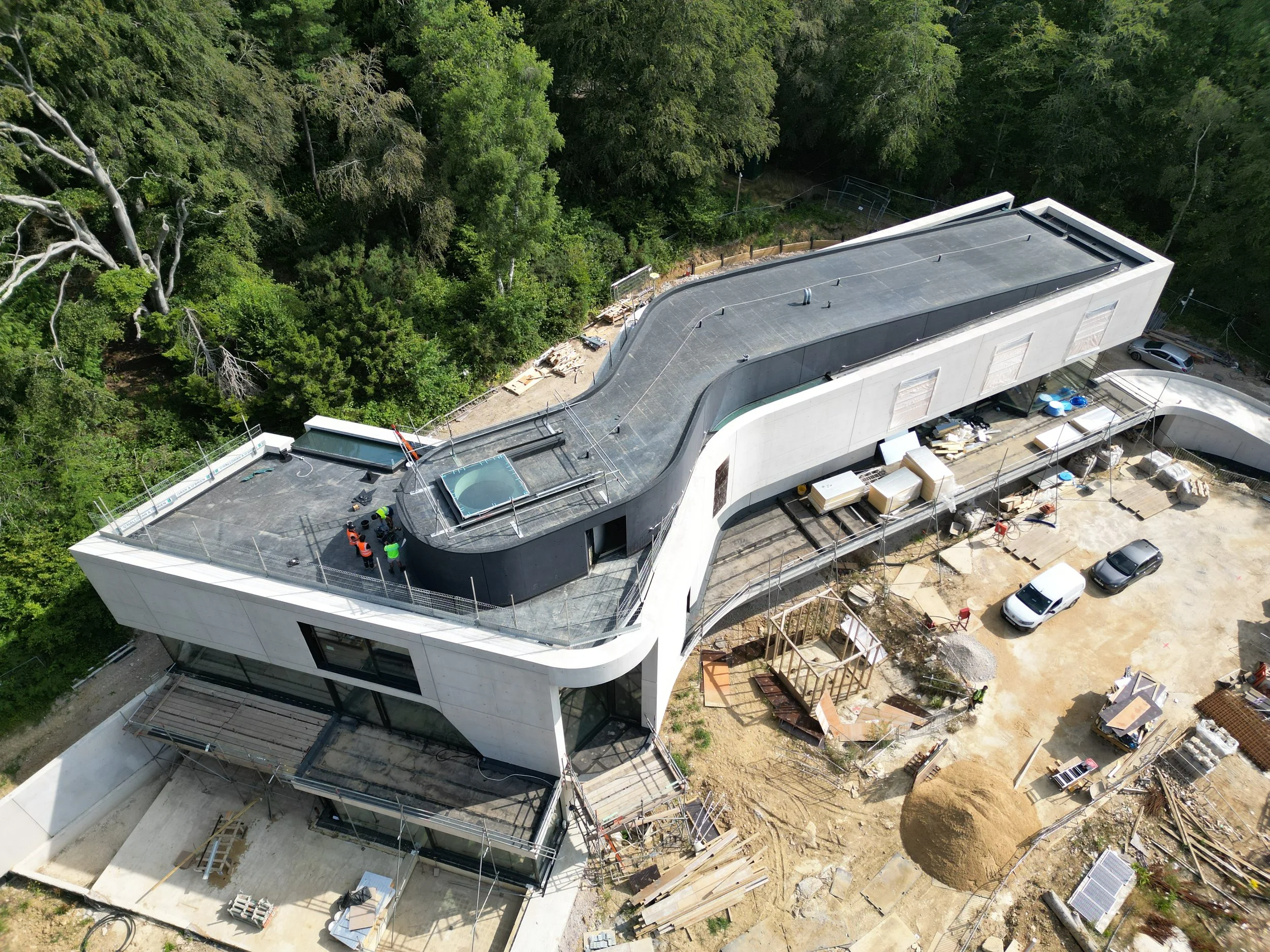 Aerial view of a modern building under construction surrounded by trees, with construction workers and materials visible on site.