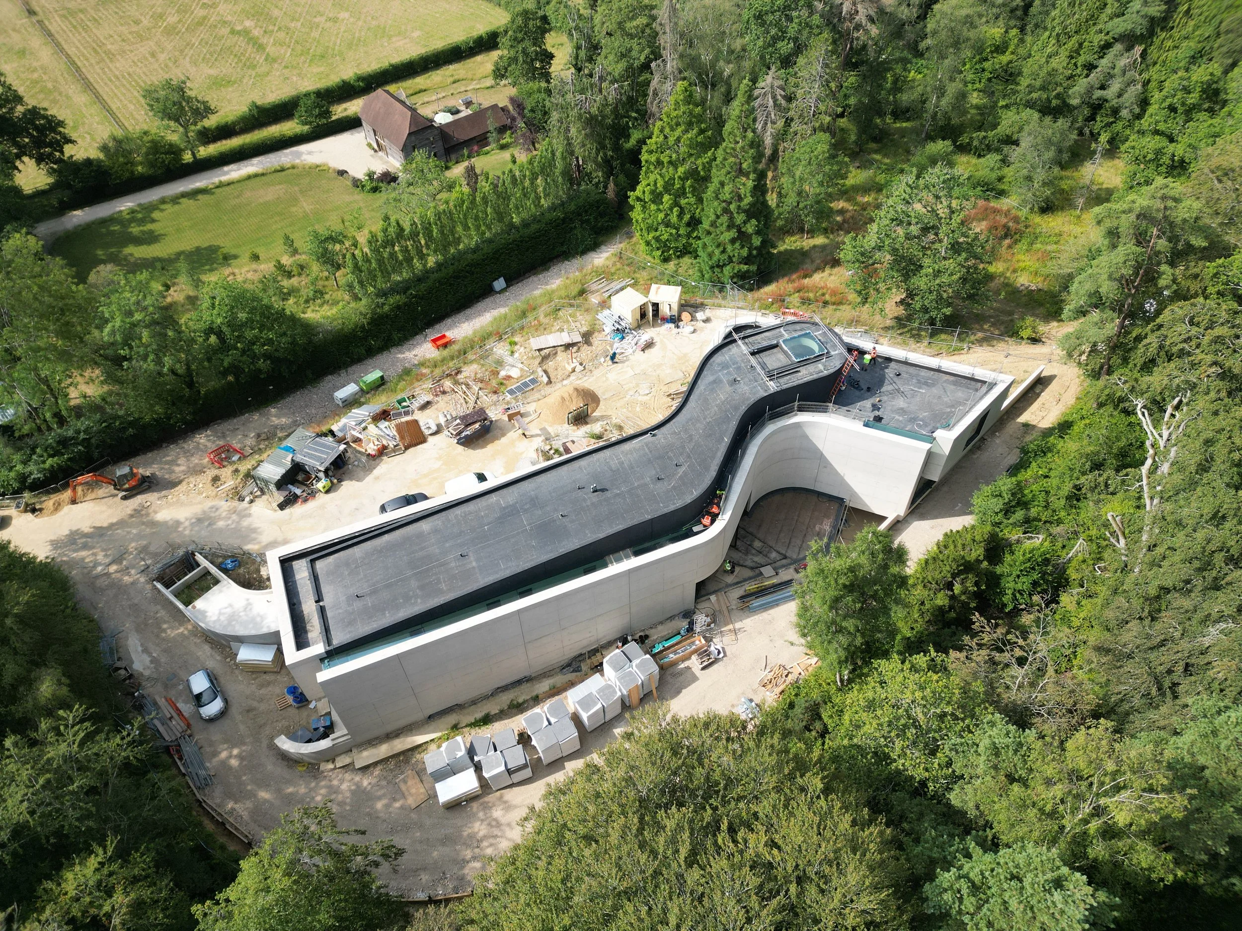 An aerial view of a construction site next to a modern white building, surrounded by lush green trees and a historic wooden house on a grassy area.