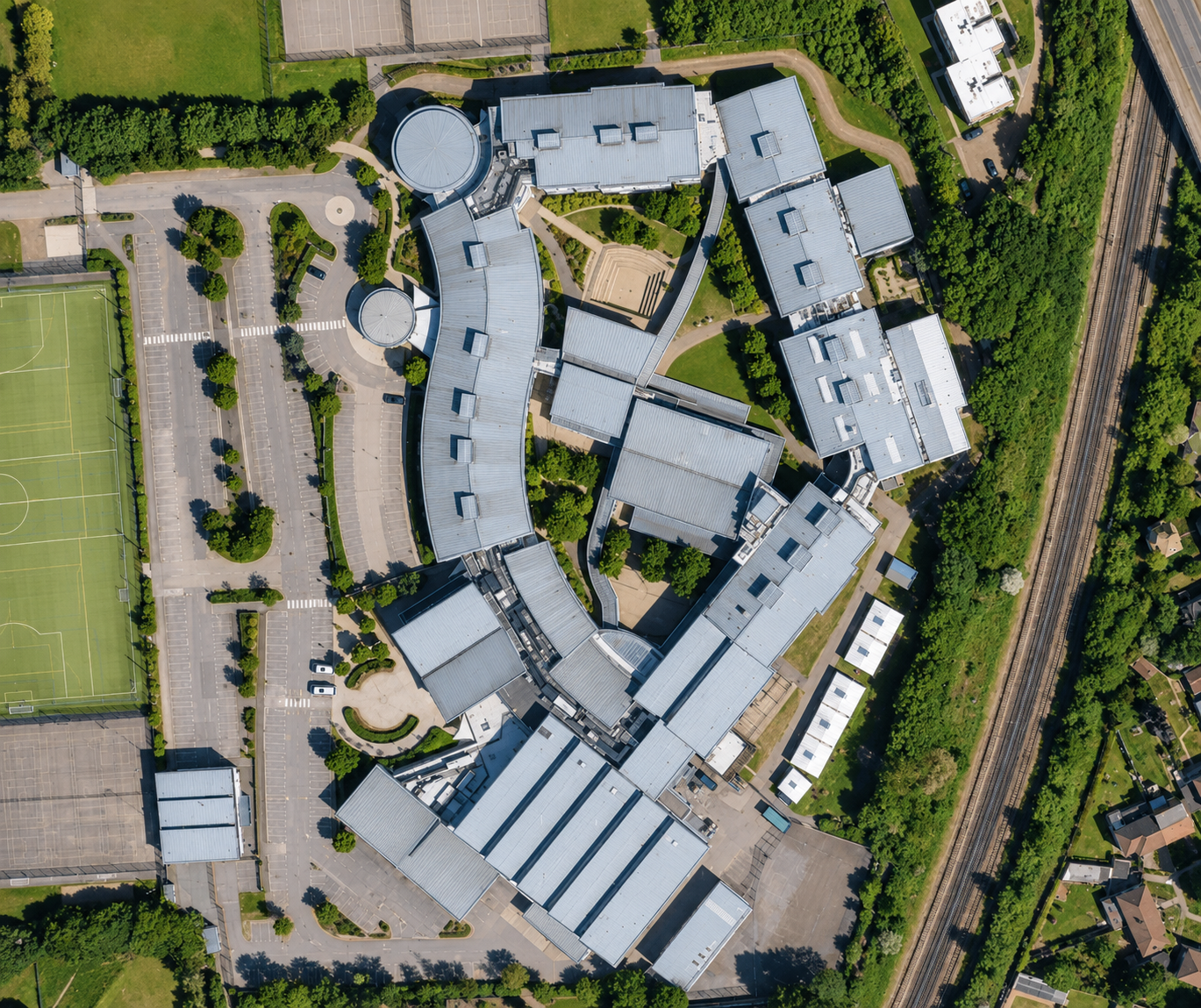 An aerial view of a modern school or university campus with curved buildings, parking lots, a sports field, and surrounding greenery.