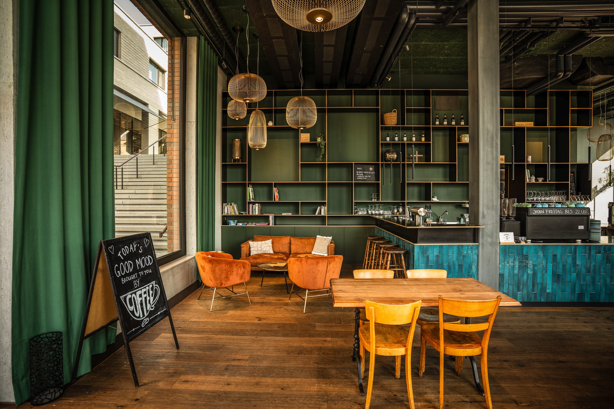Modernes Café-Interieur mit grüner Wand und Regalen, orangefarbenen Sesseln, hölzernen Tischen und Stühlen, sowie einer Tafel für Kaffeeangebote.