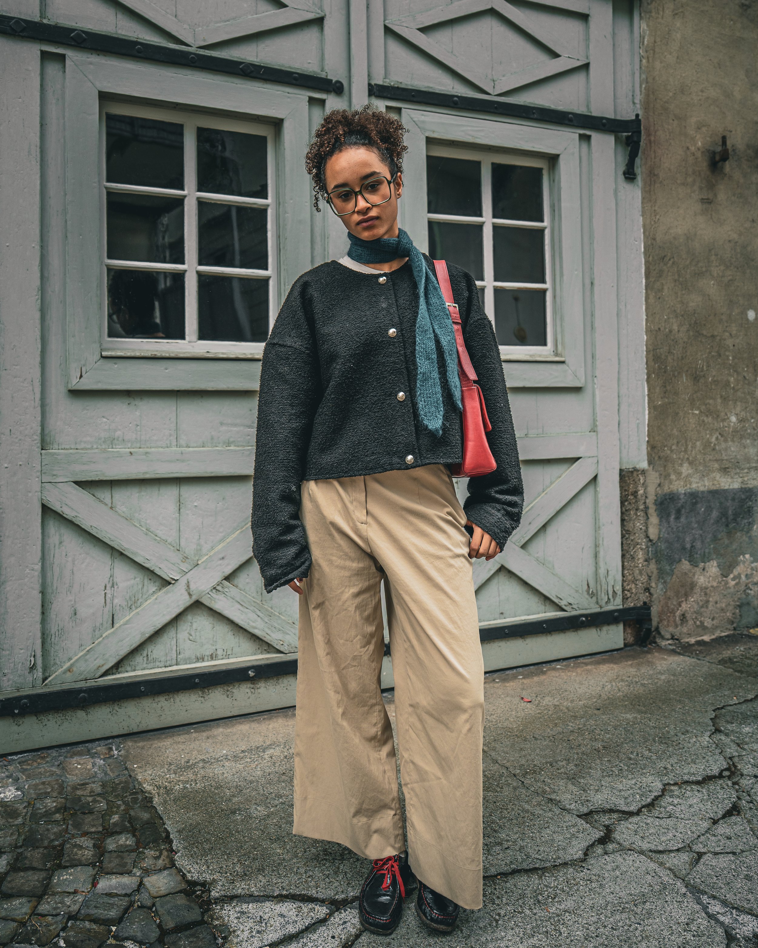 Eine Frau mit Brille trägt eine schwarze Strickjacke und beige weite Hose, posiert vor einer alten Tür.