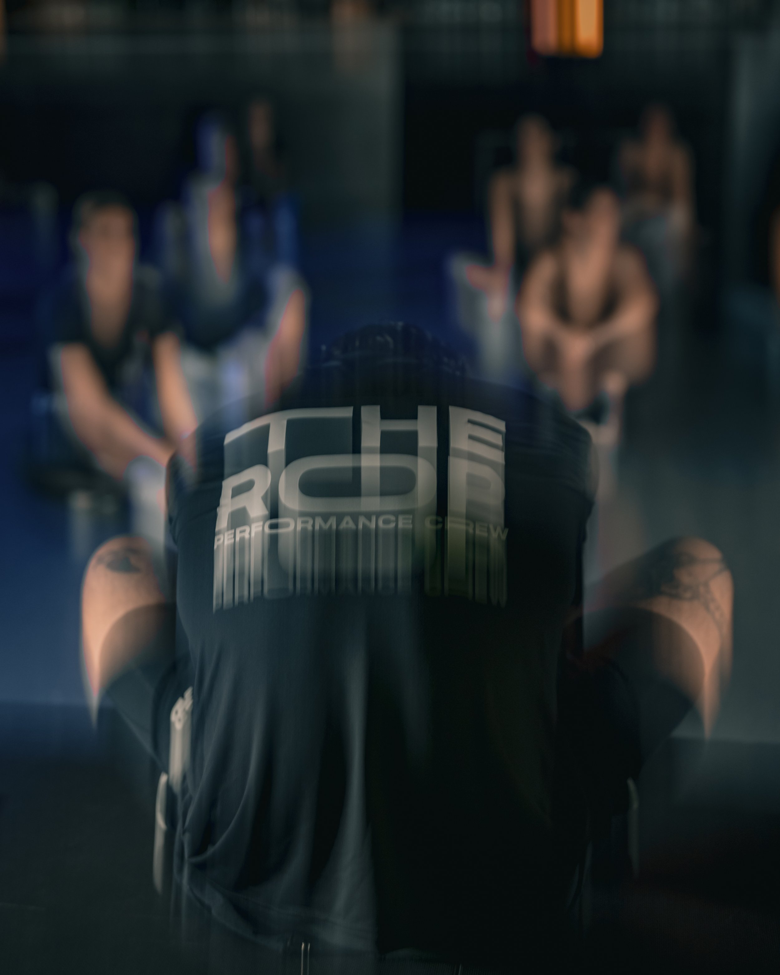 Verschwommenes Bild von Personen, die in einem Raum sitzen, mit dem Fokus auf einem Mann im Vordergrund, der ein dunkles T-Shirt mit der Aufschrift "RCR Performance Crew" trägt.
