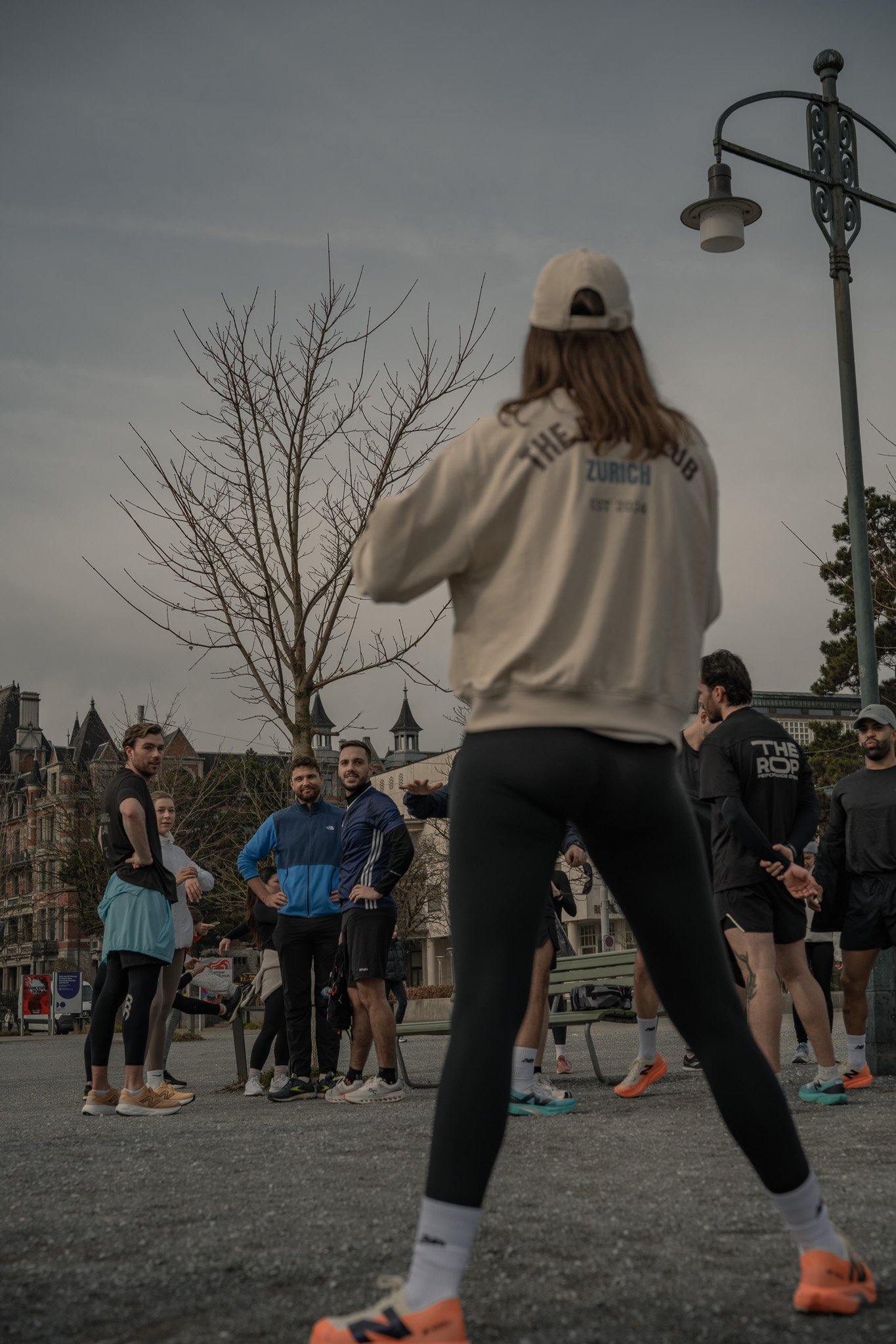 Gruppe von Menschen beim Outdoor-Training, Person mit Rückansicht in Vordergrund, Bäume und Gebäude im Hintergrund.