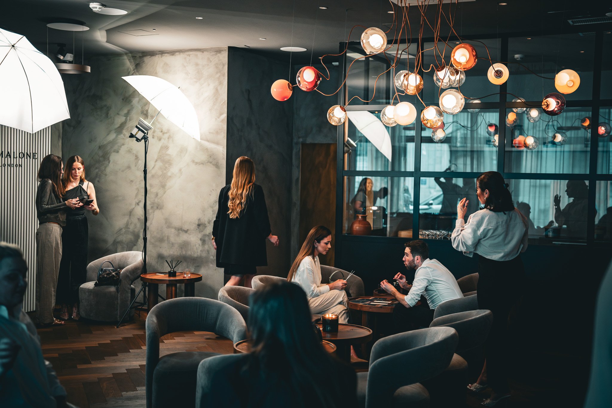 Innenraum eines stilvoll eingerichteten Cafés oder Restaurants mit mehreren Personen in Gesprächssituationen, modernen Möbeln und einer markanten Hängelampe.