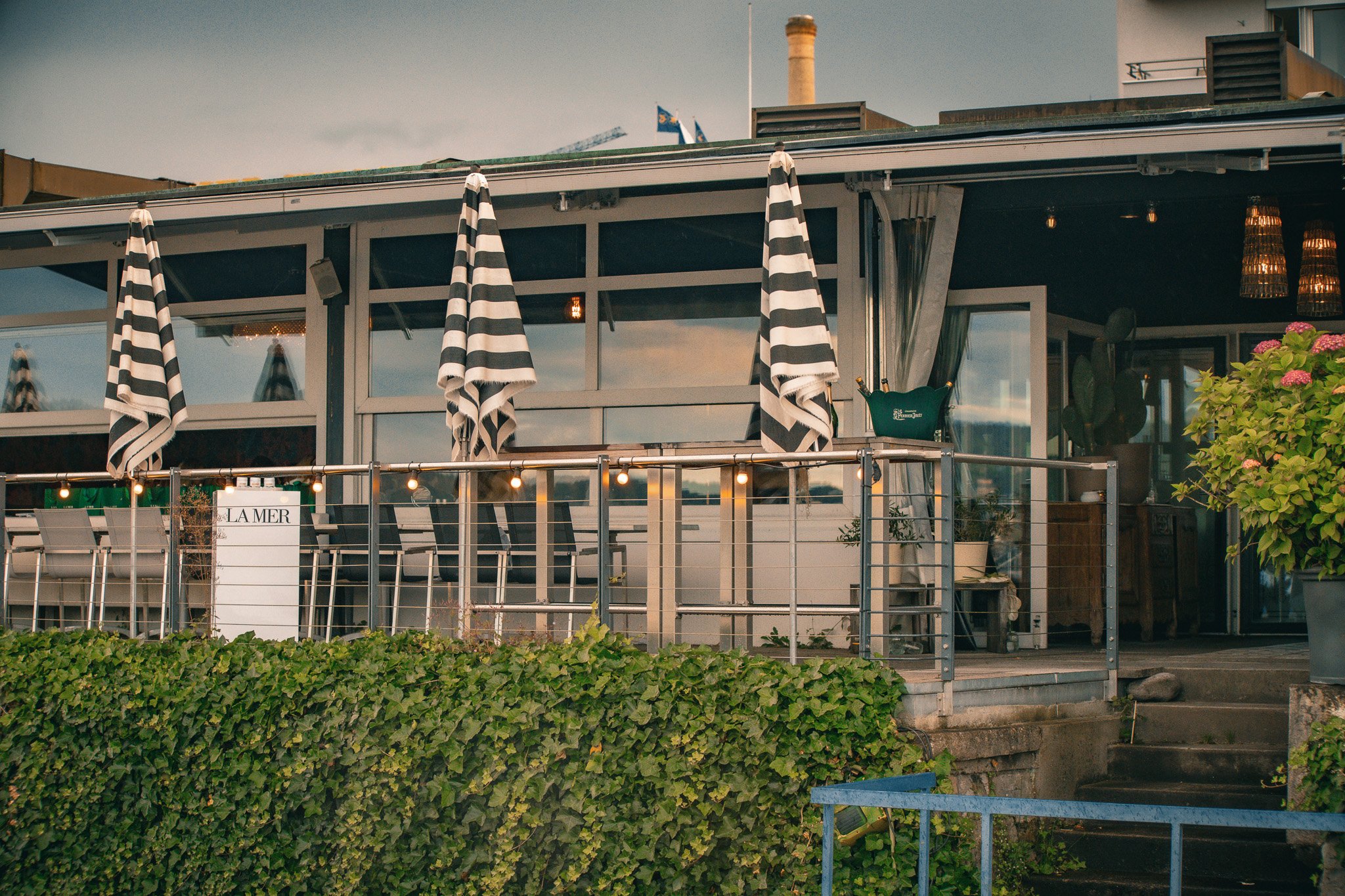 Außenbereich eines modernen Cafés mit weißen Tischen, gestreiften Sonnenschirmen und Glasfenstern, umgeben von Pflanzen.