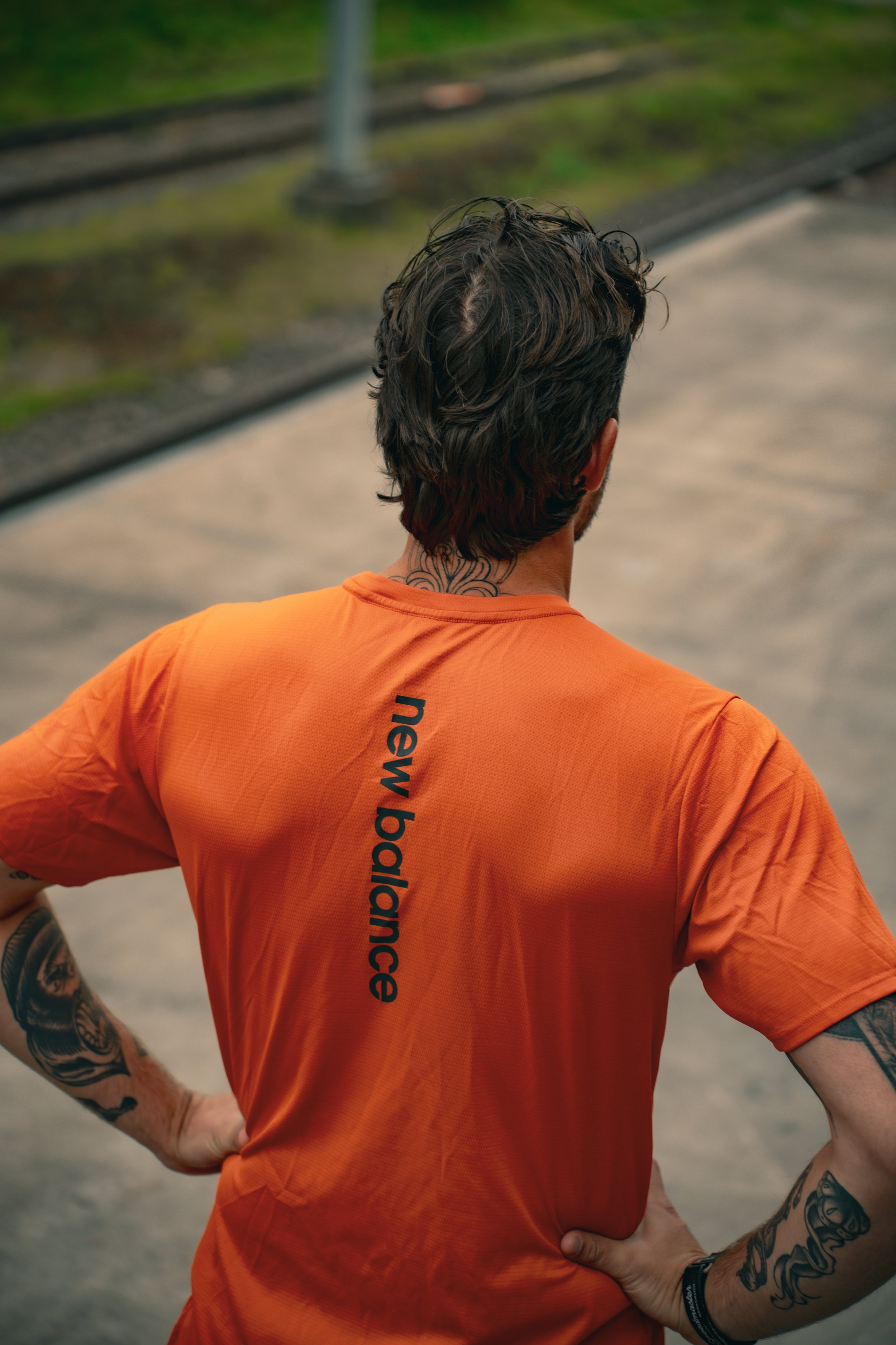 Rückansicht einer Person in einem orangefarbenen "New Balance"-Shirt auf einem Bahnsteig, mit Tätowierungen an den Armen.