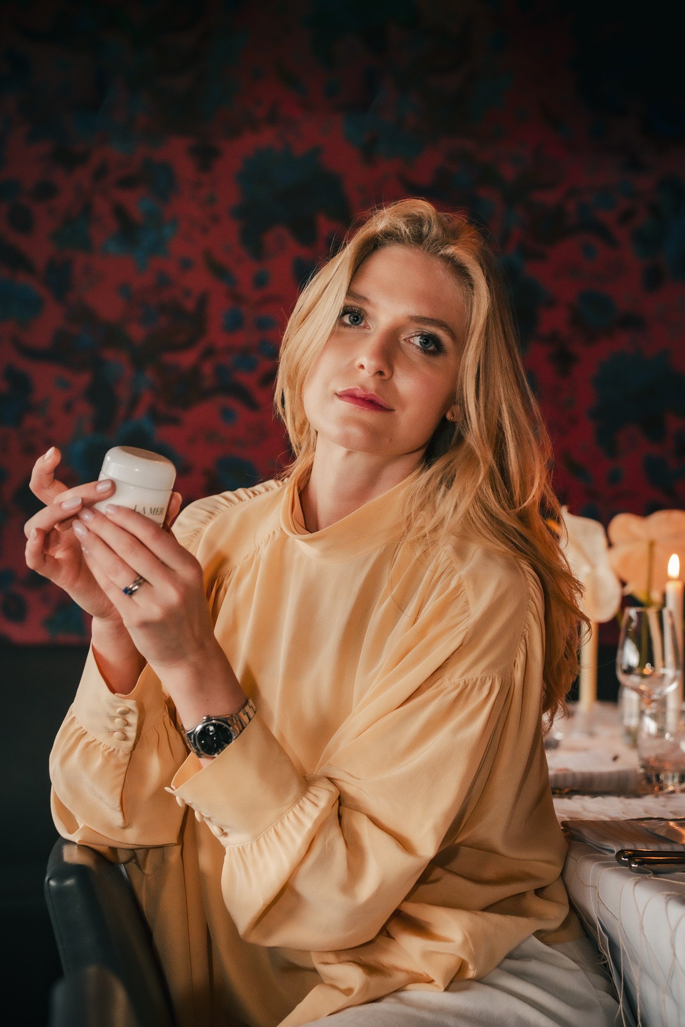 Frau in gelber Bluse hält ein Kosmetikprodukt in der Hand, sitzt an einem Tisch, Kerze im Hintergrund.