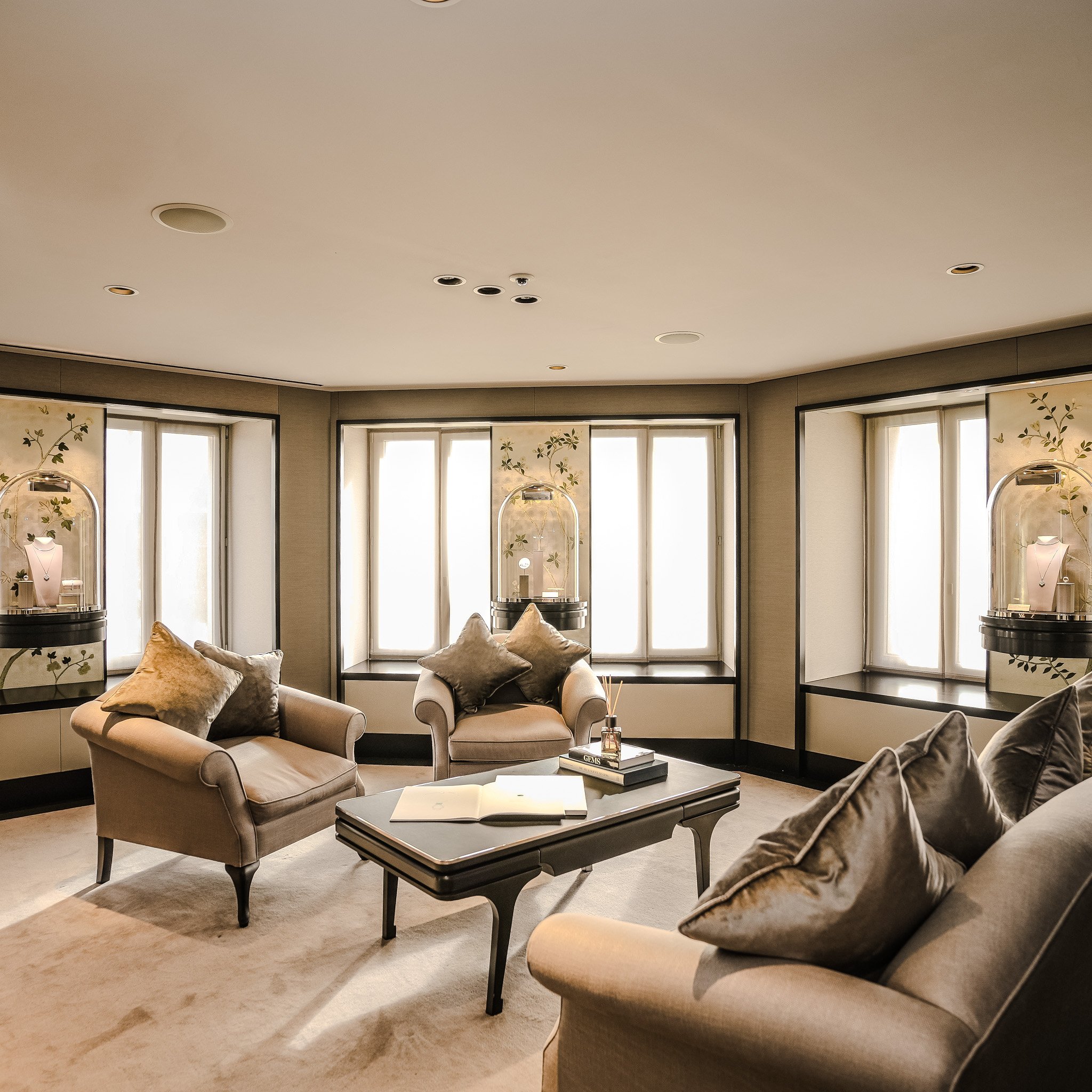 Luxuriöses Wohnzimmer mit beigen Sesseln, einem Tisch mit Büchern, großen Fenstern und dekorativen Vitrinen mit Schmuck.