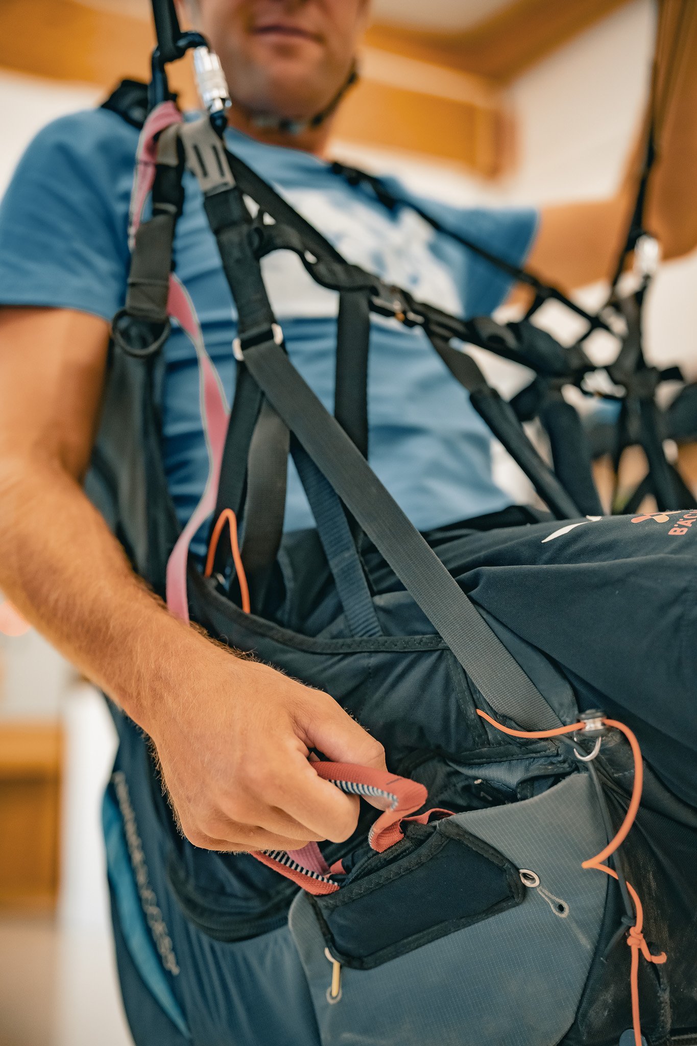Nahaufnahme eines Mannes, der ein Paragliding-Gurtzeug trägt und es zu justieren scheint. Der Mann trägt ein blaues T-Shirt und greift nach einem roten Gurtband. Der Hintergrund ist unscharf.