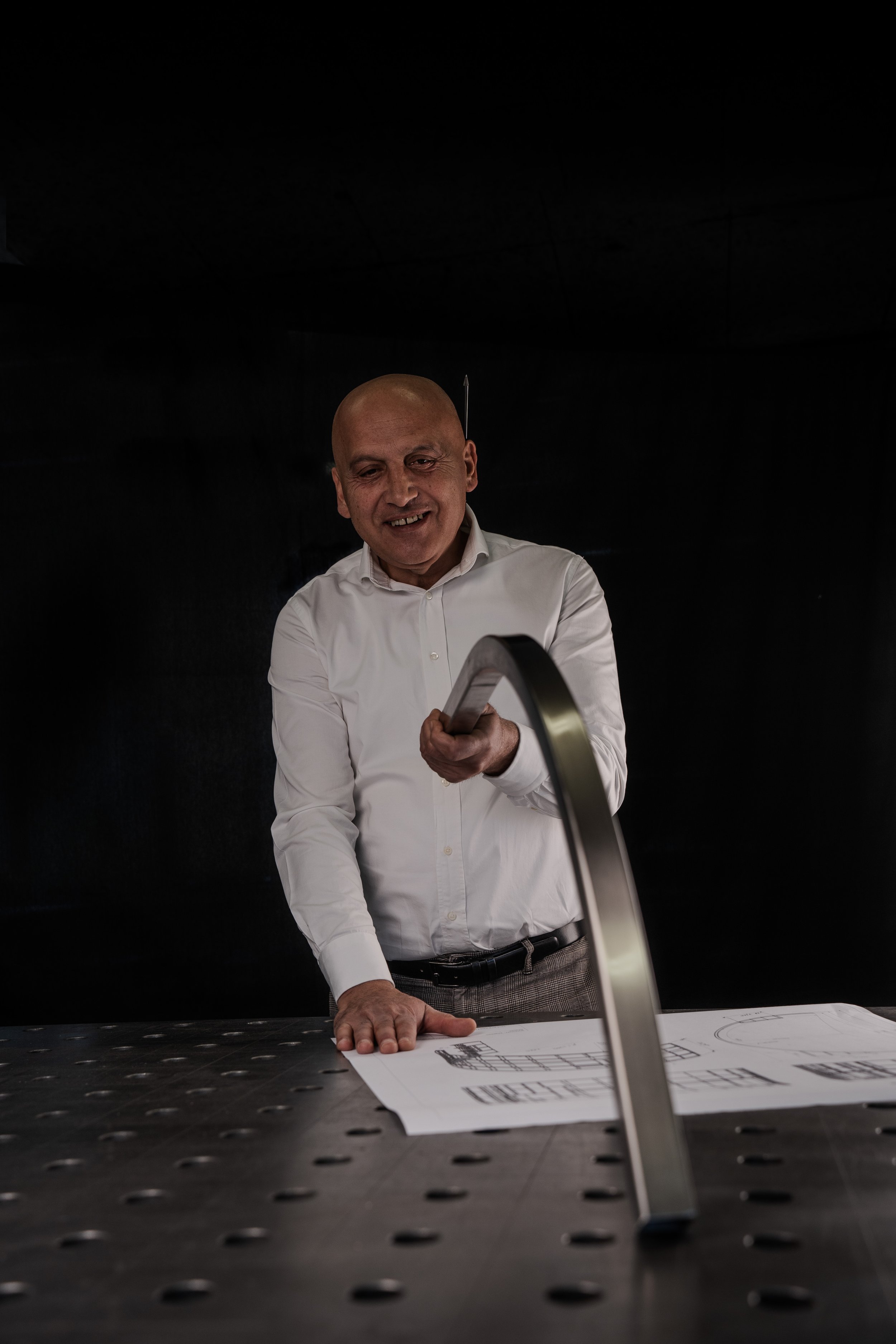 Mann in weißem Hemd mit einem gebogenen Metallstück und technischen Zeichnungen auf einem Tisch.