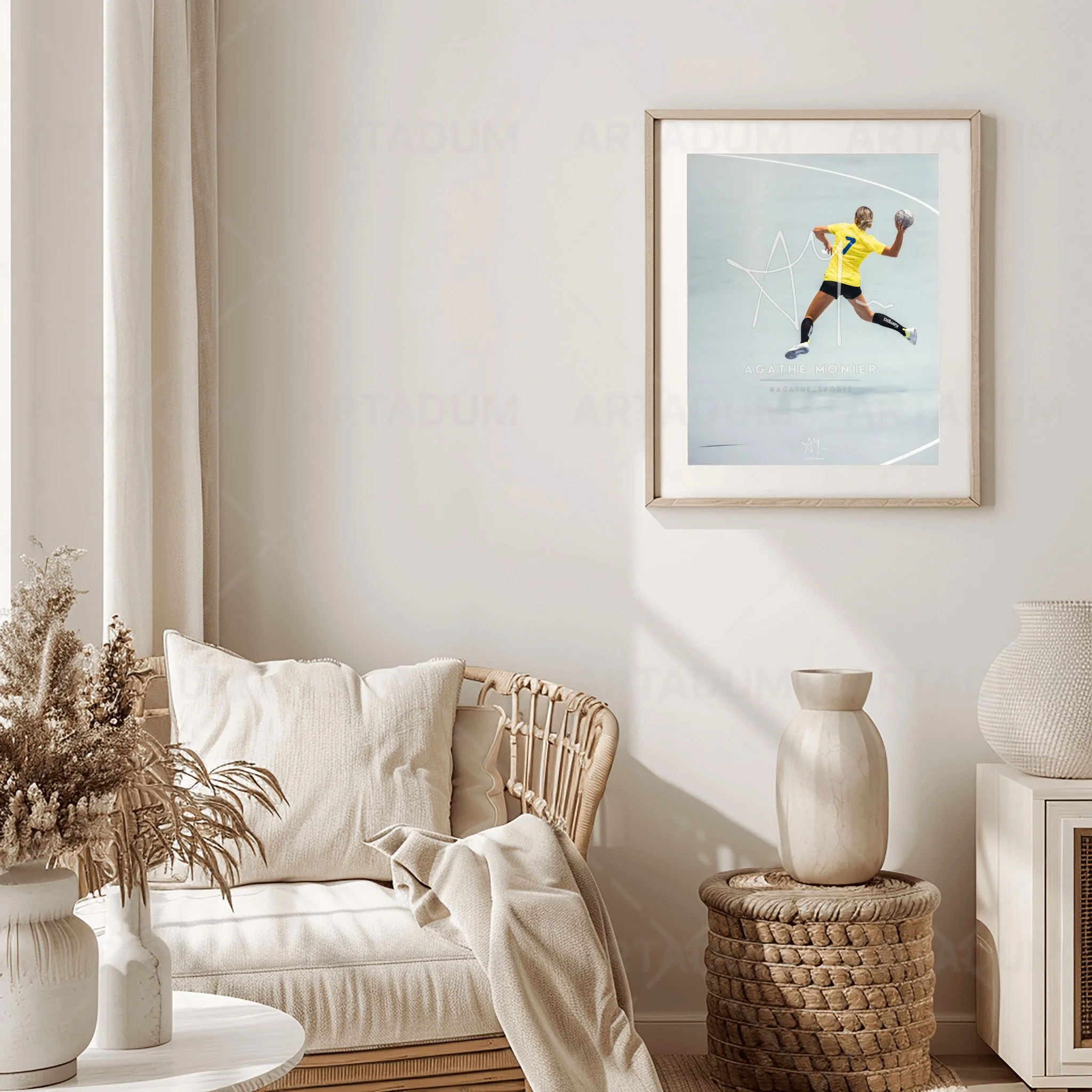 Une pièce décorée dans un style minimaliste avec un canapé beige, des coussins blancs, une plante séchée dans un vase blanc, et un tableau avec une personne portant un maillot jaune en mouvement. Un vase beige sur un support en rotin.