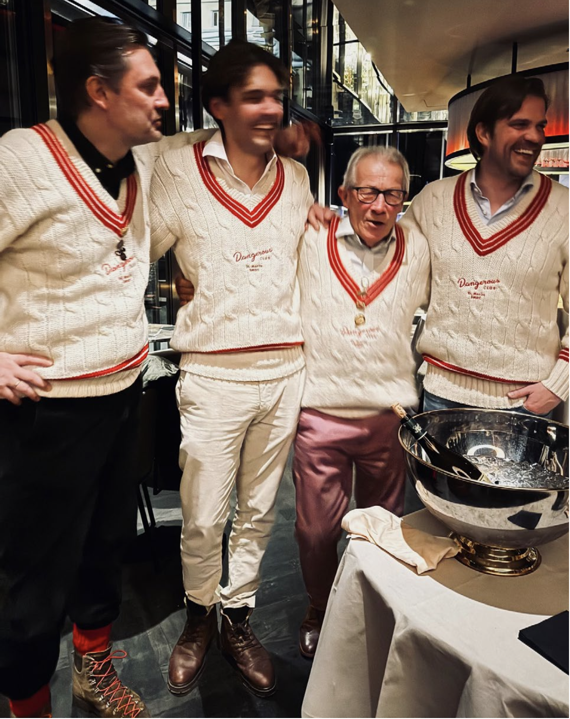Four men in cream-colored sweaters with red and white knit patterns, standing together at a celebration, smiling and laughing. A bucket with a bottle is in front of them.