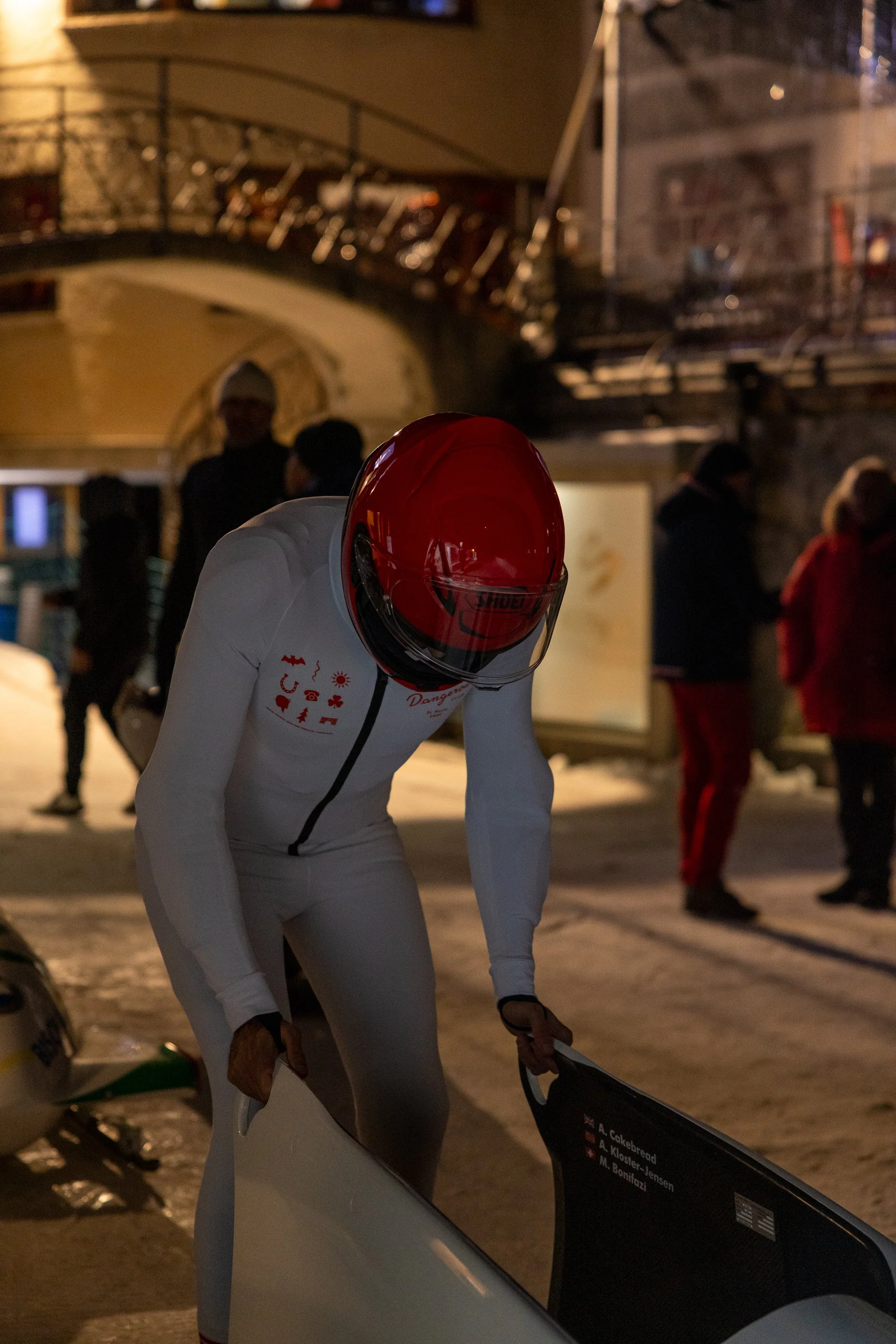 Person dressed in a white cycling suit with a red helmet leaning over, with a group of people in the background at night in an outdoor setting.