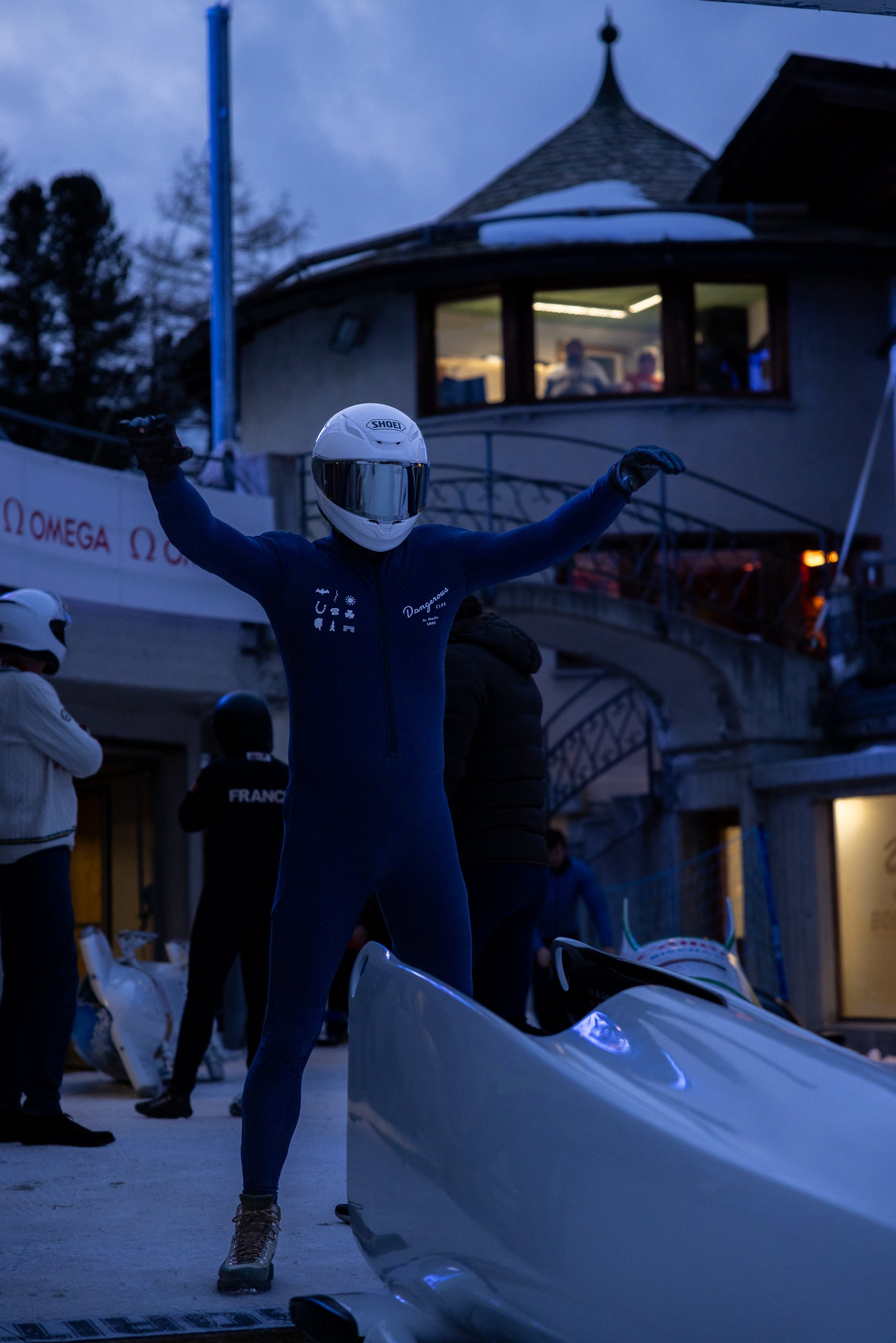 A person dressed in a blue speed skating suit and a white helmet strikes a victory pose at dusk in front of a white racing vehicle. There are spectators and a building with large windows and a pointed roof in the background.