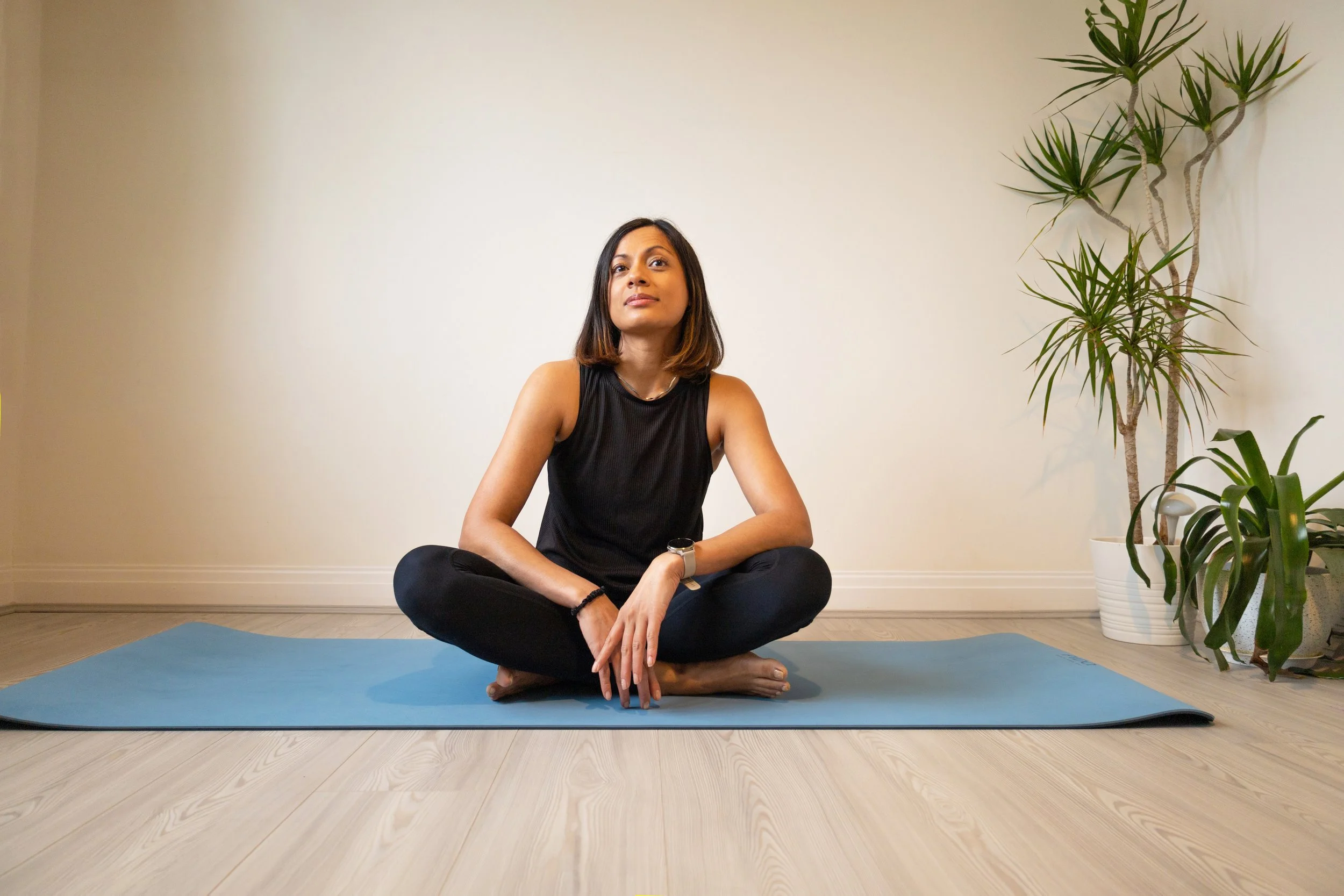 A woman sitting cross-legged on a yoga mat in a room with wooden floors and a plain white wall, with large potted plants nearby.
