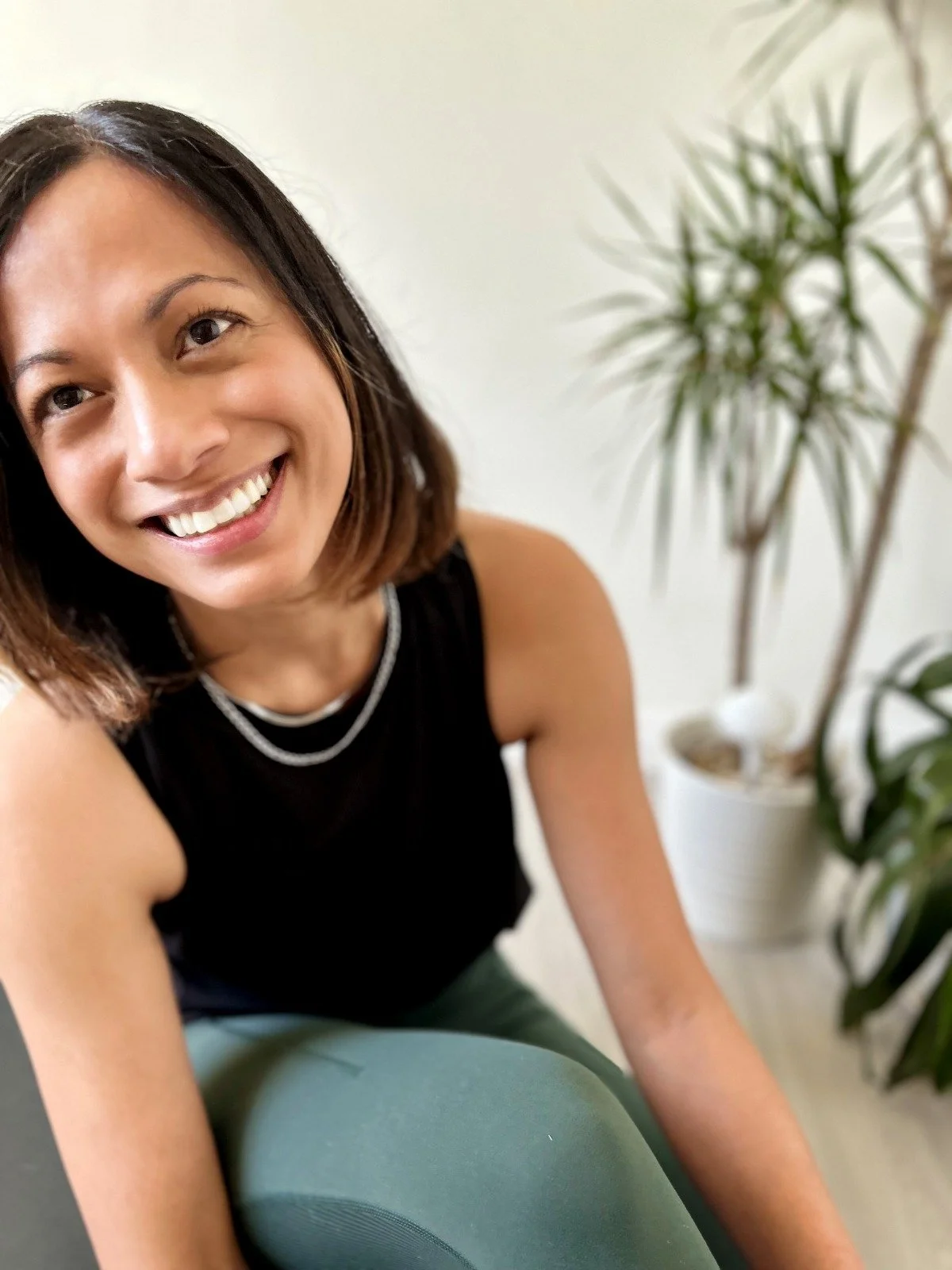 A smiling woman with shoulder-length brown hair wearing a black sleeveless top and layered necklaces, sitting near houseplants on a white background.