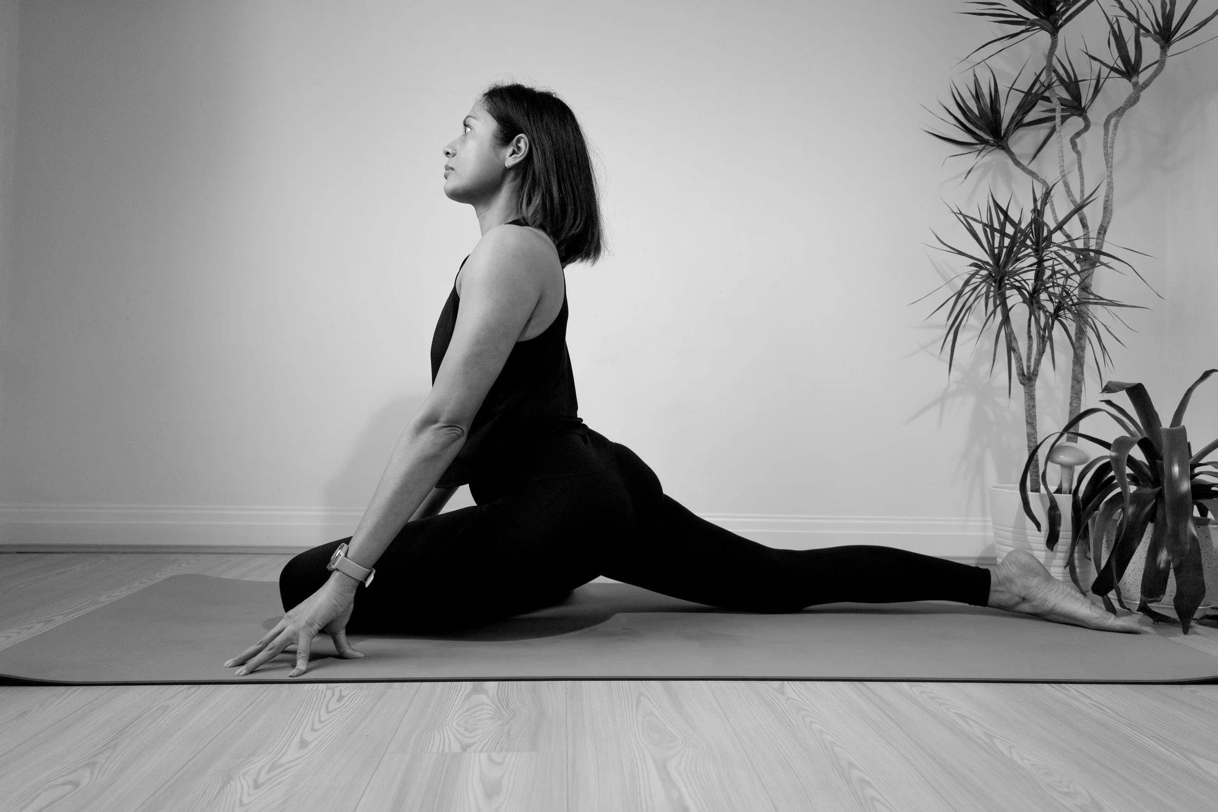 A woman practicing yoga in a lunge position on a yoga mat indoors. She is wearing black workout clothes and a watch, with her left hand on the floor and her right arm resting on her right knee. Behind her, there is a large potted plant with tall, spiky leaves against a plain wall.