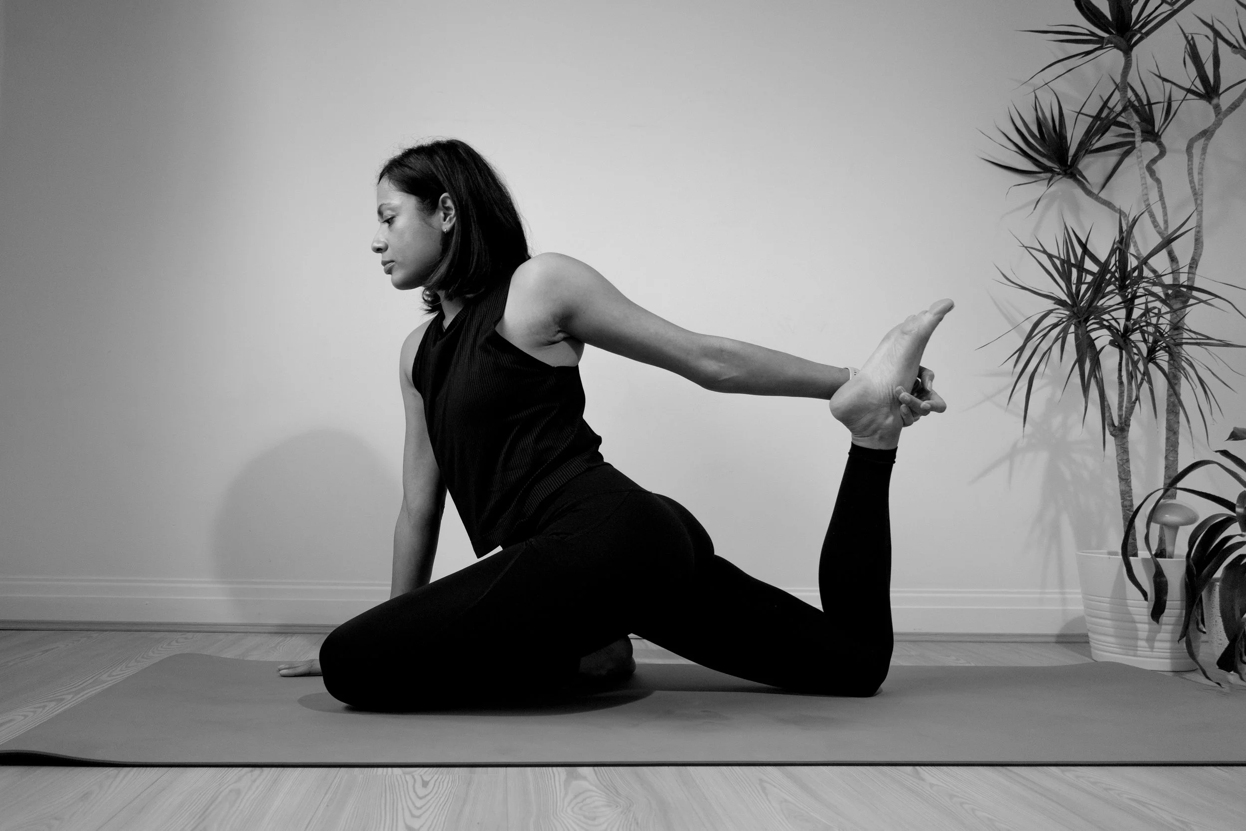 A woman practicing yoga indoors, executing a modified pigeon pose, wearing a black sleeveless top and black leggings, with a potted plant nearby.