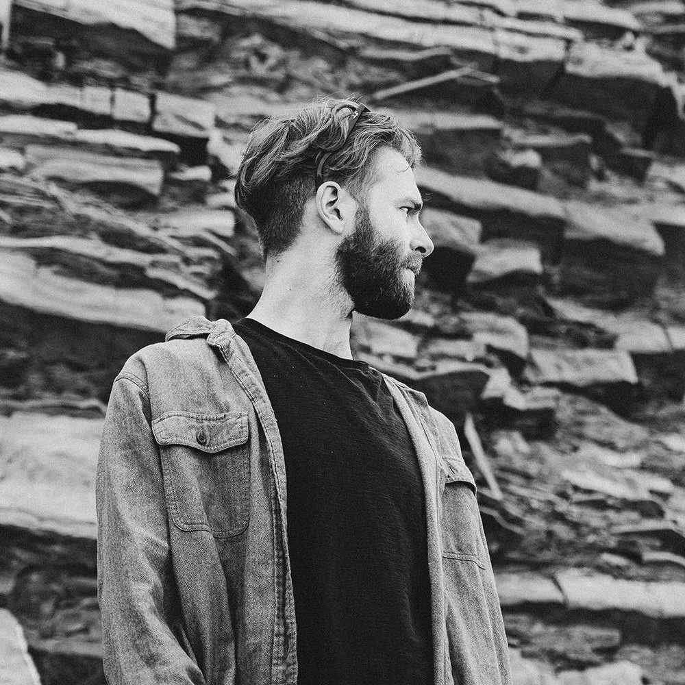Black and white photo of a man with a beard wearing a denim jacket and black shirt, standing in front of a rocky background.