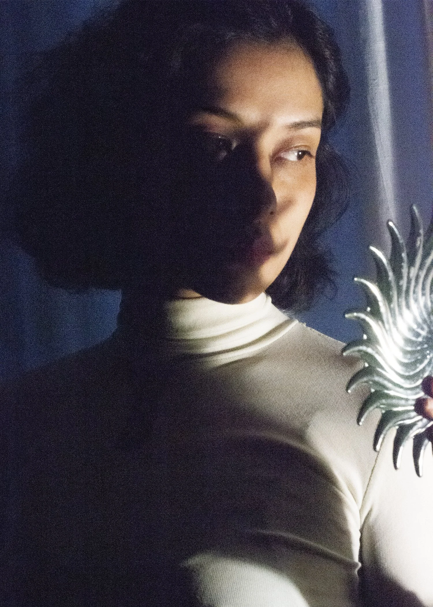 Person with short hair standing in dim light, holding a decorative silver mirror shaped like sun rays.
