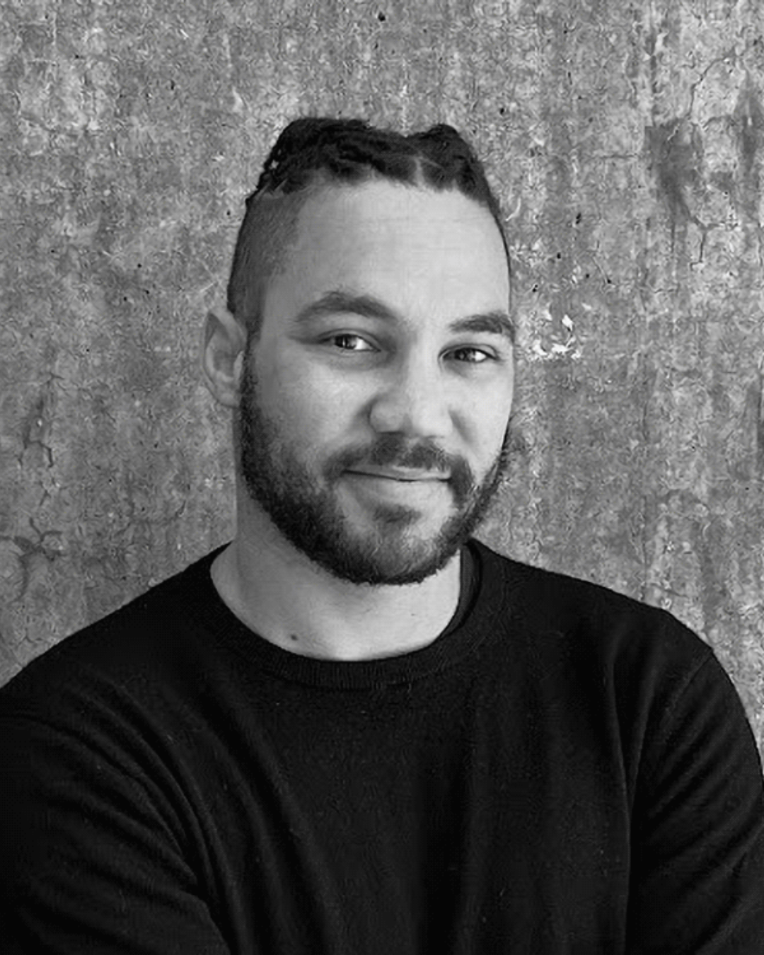 Black and white photo of a man with braided hair and a beard, wearing a black sweater, smiling slightly, against a textured concrete wall.
