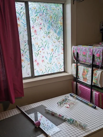 A cutting table in front of a large window. The window is covered in a cling that looks like floral stained glass. Next to the table is several shelves full of bolts of colorful fabric.